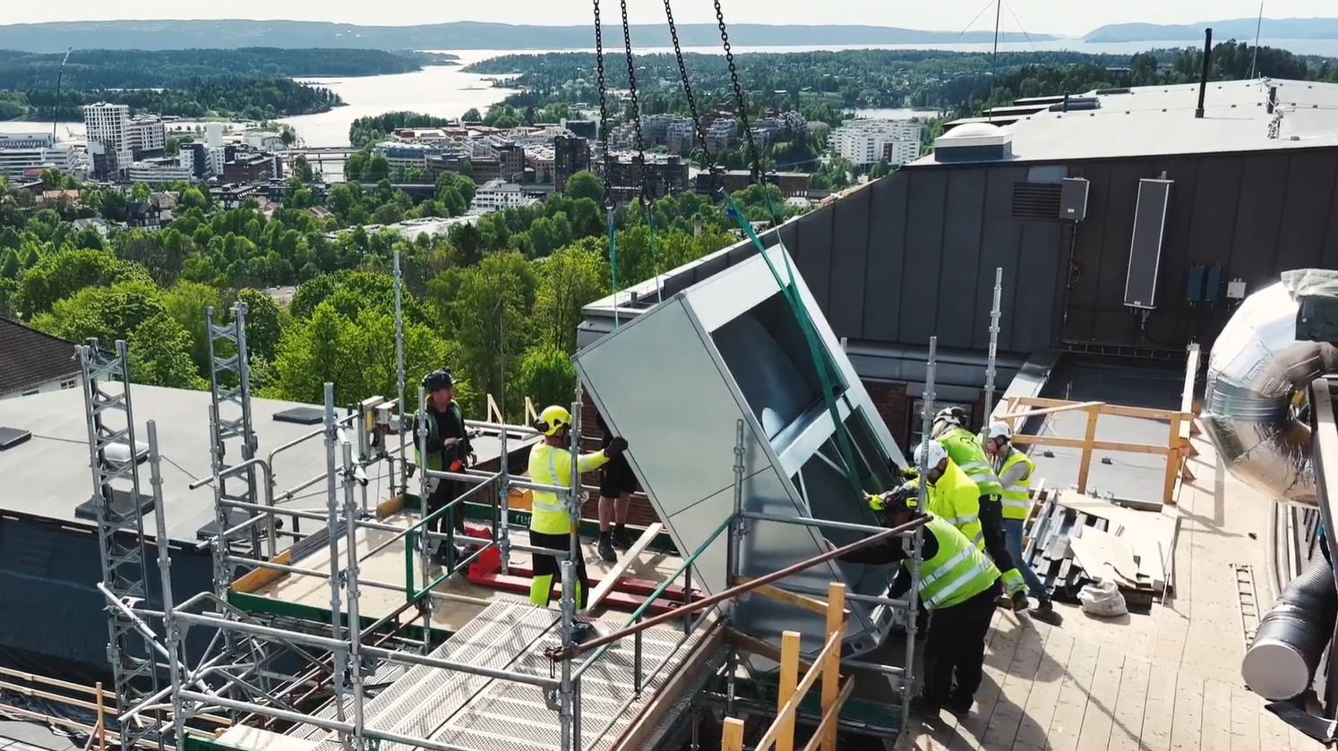 Seks arbeidere i verneutstyr står på taket på Bærum sykehus og på tilhørende stilas og samarbeider om å ta i mot eller løfte av en del av et nytt aggregat. Aggregatdelen har blitt løftet opp på taket av en mobilkran og er sikret med kjetting og belter. Utsikt utover Sandvika og Oslofjorden i bakgrunnen.