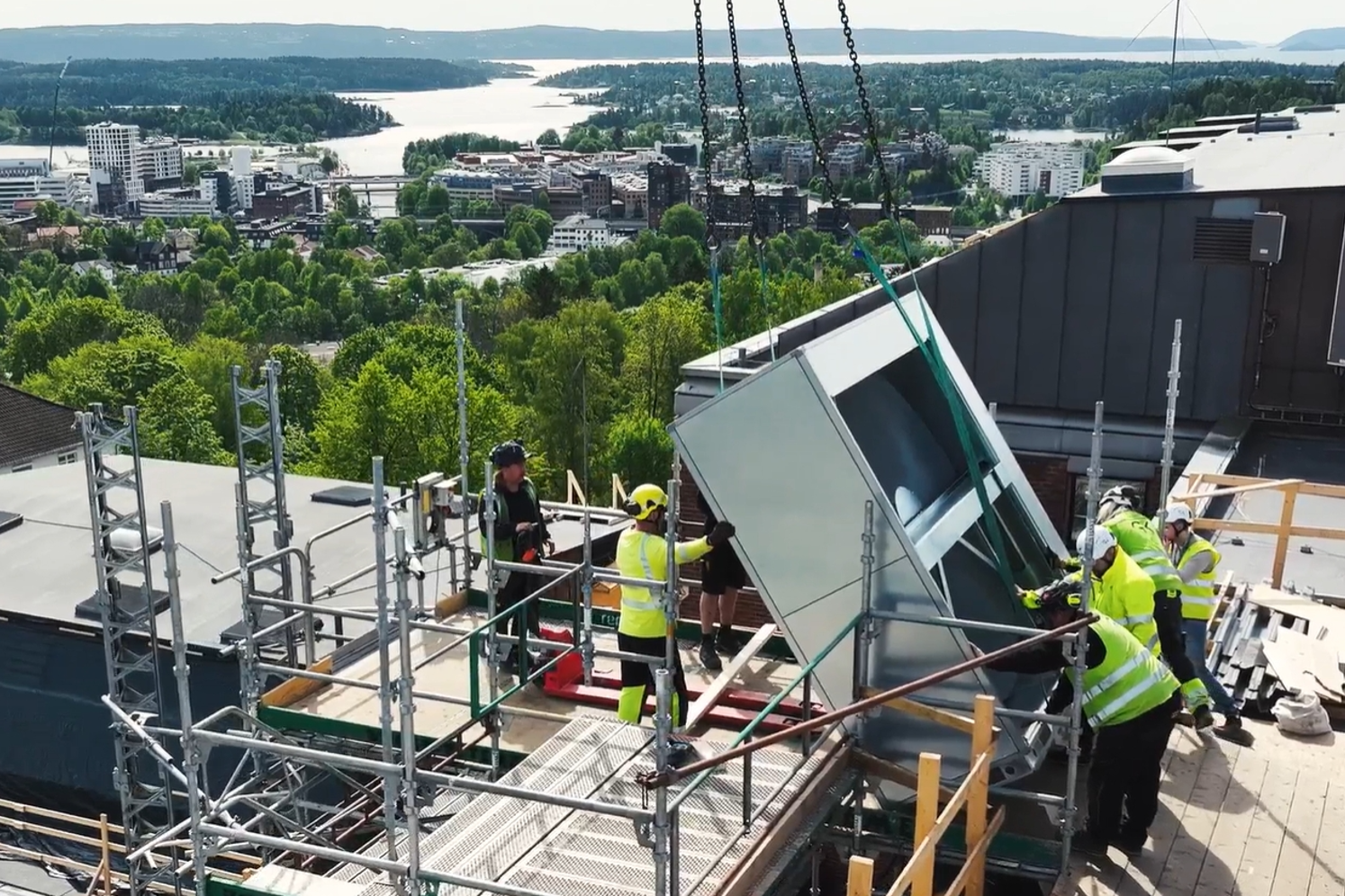 Seks arbeidere i verneutstyr står på taket på Bærum sykehus og på tilhørende stilas og samarbeider om å ta i mot eller løfte av en del av et nytt aggregat. Aggregatdelen har blitt løftet opp på taket av en mobilkran og er sikret med kjetting og belter. Utsikt utover Sandvika og Oslofjorden i bakgrunnen.