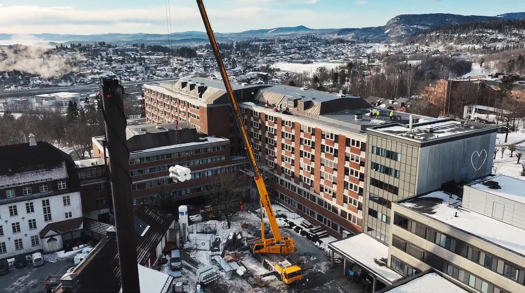Orange mobilkran som står i bakgården utenfor Bærum sykehus. Kranen heiser opp utstyr. Ser fasaden til sykehuset bak kranen og utsikt utover Sandvika mot Slependen.