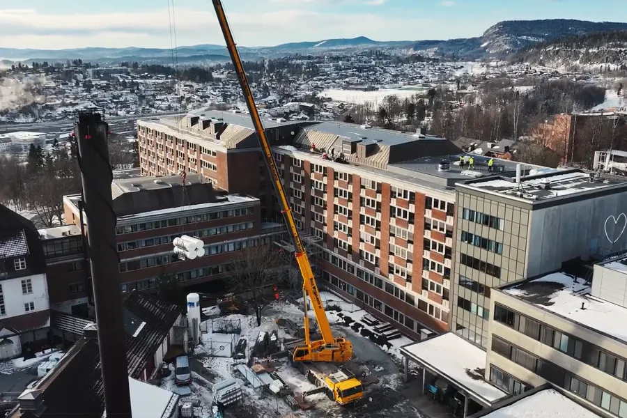 Orange mobilkran som står i bakgården utenfor Bærum sykehus. Kranen heiser opp utstyr. Ser fasaden til sykehuset bak kranen og utsikt utover Sandvika mot Slependen.