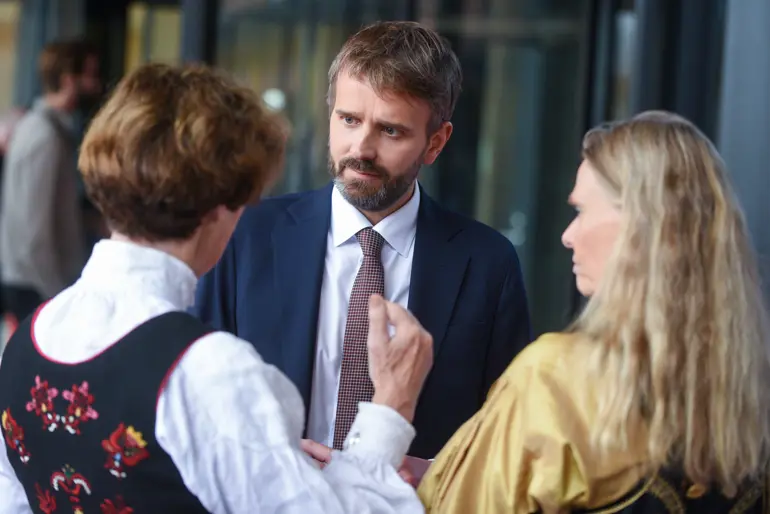 Helse- og omsorgsminister Jan Christian Vestre ser og lytter til styreleder i Vestre Viken, Siri Hatlen. Hatlen står til venstre med bakhodet til, og er iført sort bunad og hvit bunadsskjorte, og gestikulerer med høyrehånden. Til høyre for Hatlen står administrerende direktør i Vestre Viken, Lisbeth Sommervoll, også iført sort bunad med gul bunadsskjorte.