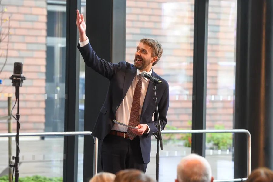 Helse- og omsorgsminister Jan Christian Vestre holdt tale foran oppmøtte under den offisielle åpningen av Drammen sykehus høsten 2025. Vestre er pent kledd i dress, hvit skorte og slips, står på en scene inne i foajeen til sykehuset med store vinduer bak seg. En mikrofon står foran han og man kan så vidt se bakhodene til noen av de oppmøtte. Vestre holder høyre hånden opp i luften og ser opp til høyre. Han holder talekort i venstre hånden.