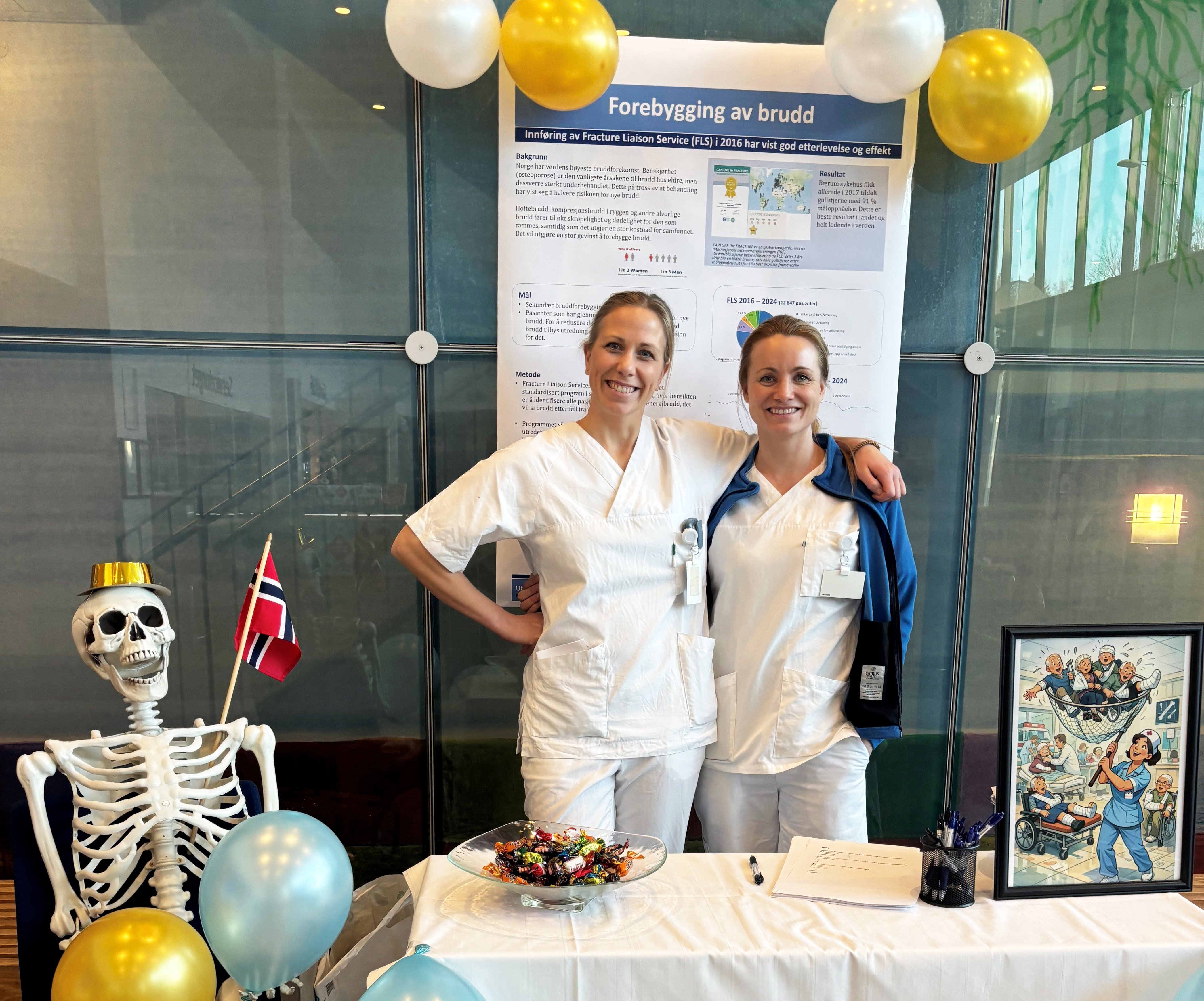 To sykepleiere i hvit uniform. De står oppstilt og smiler bak et bord med hvit duk. På duken står det en skål med tvist, et ark med noe tekst, penner og et innrammet bilde av en sykepleier som har fanget bruddpasienter med en håv. Til venstre sitter et plastskjelett som er pyntet med festhatt og flagg, og gull og blå ballonger. Bak sykepleierne er det en glassvegg og en plakat hvor det står om avdelingens erfaringer med forebygging av brudd. Det er festet fire gull og hvite ballonger over plakaten.