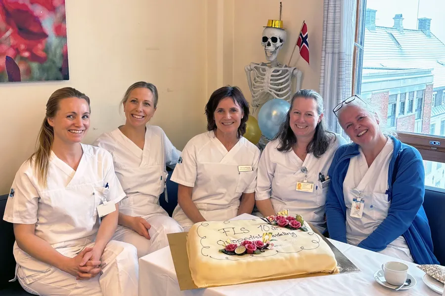 En gruppe sykepleiere og en ortoped feirer med marsipankake. De sitter på en svart sofa i hvit arbeidsuniform med en marsipankake med teksten "FLS 10 år Ortopedisk avdeling" på bordet foran seg. De smiler til kamera. Det er et vindu bak de med utsikt utover fasaden til en sykehusfløy og et plastskjelett som er pyntet med flagg og festhatt.