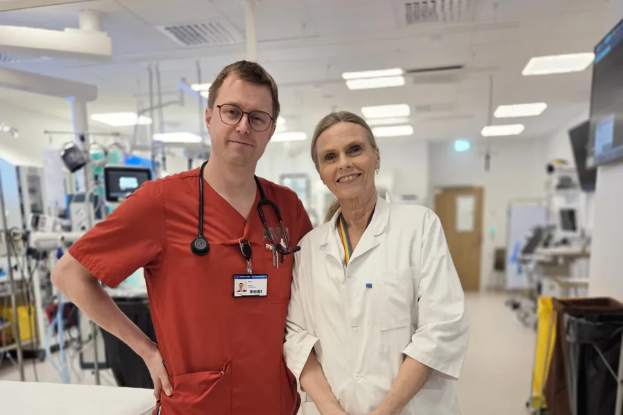 To helsepersonell i uniform som poserer på et bilde. Mannlig helsepersonell til venstre er iført en rød sykehusskjorte, har stetoskop rundt halsen og synlig id-kort. Kvinnelig helsepersonell er iført hvit legefrakk. Bak de ser man konturene av akuttmedisinsk utstyr, skjermer, søppelbøtter, traller og sykehusseng.