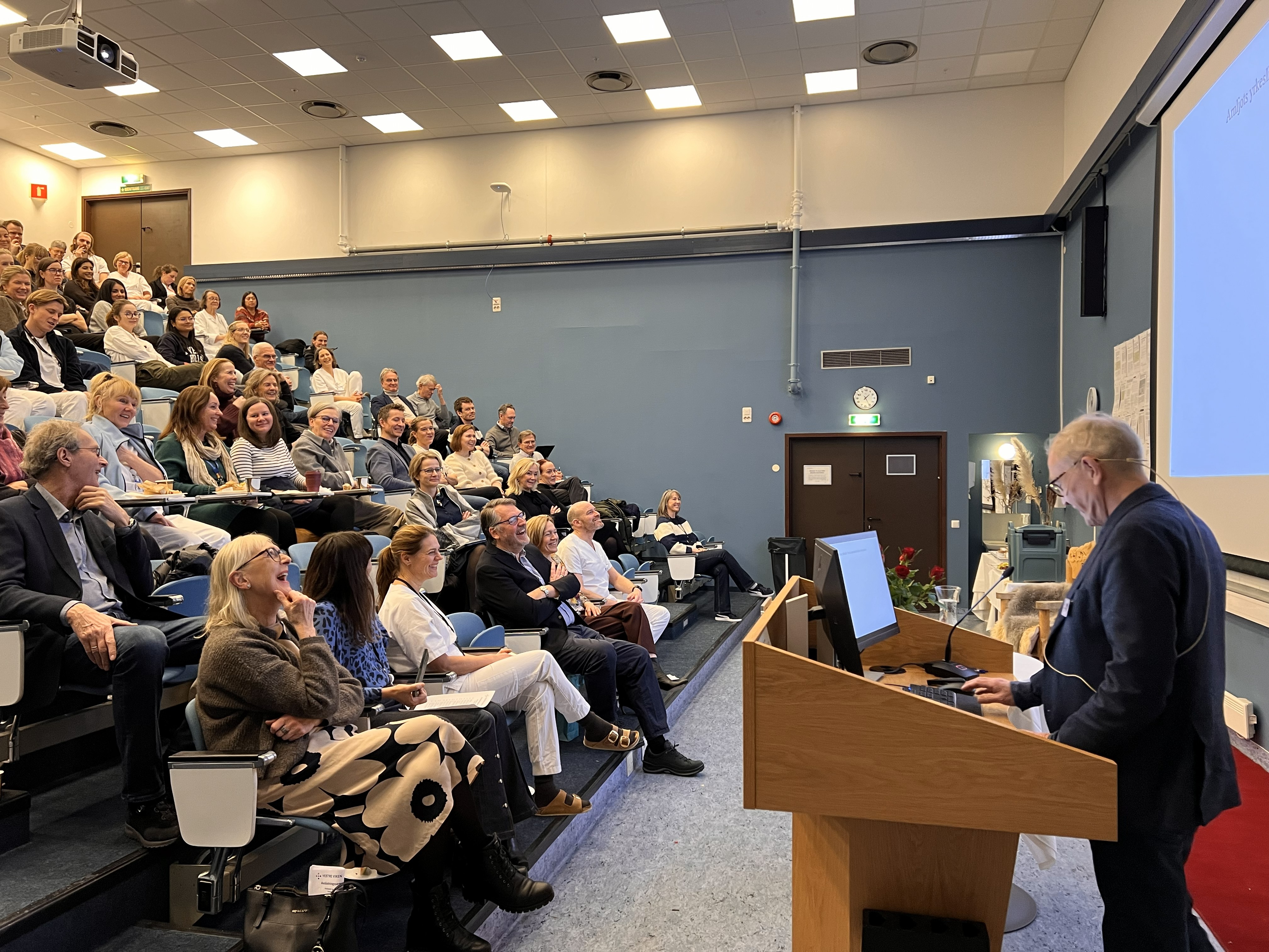 Tekst generert av copilot: Et auditorium fylt med tilhørere som følger en presentasjon. Foredragsholder står ved pulten foran lerretet, og rommet har tydelig preg av faglig samling og konferanse.