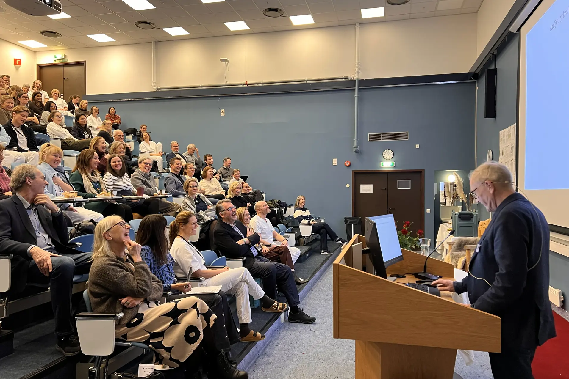 Tekst generert av copilot: Et auditorium fylt med tilhørere som følger en presentasjon. Foredragsholder står ved pulten foran lerretet, og rommet har tydelig preg av faglig samling og konferanse.