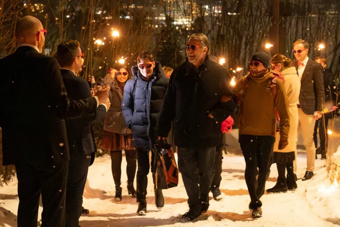 Tekst generert av copilot: En gruppe personer går sammen utendørs i vintermørket, ikledd mørke dresser og yttertøy, mens de holder fakler. Snø på bakken og lys fra faklene skaper en høytidelig stemning.