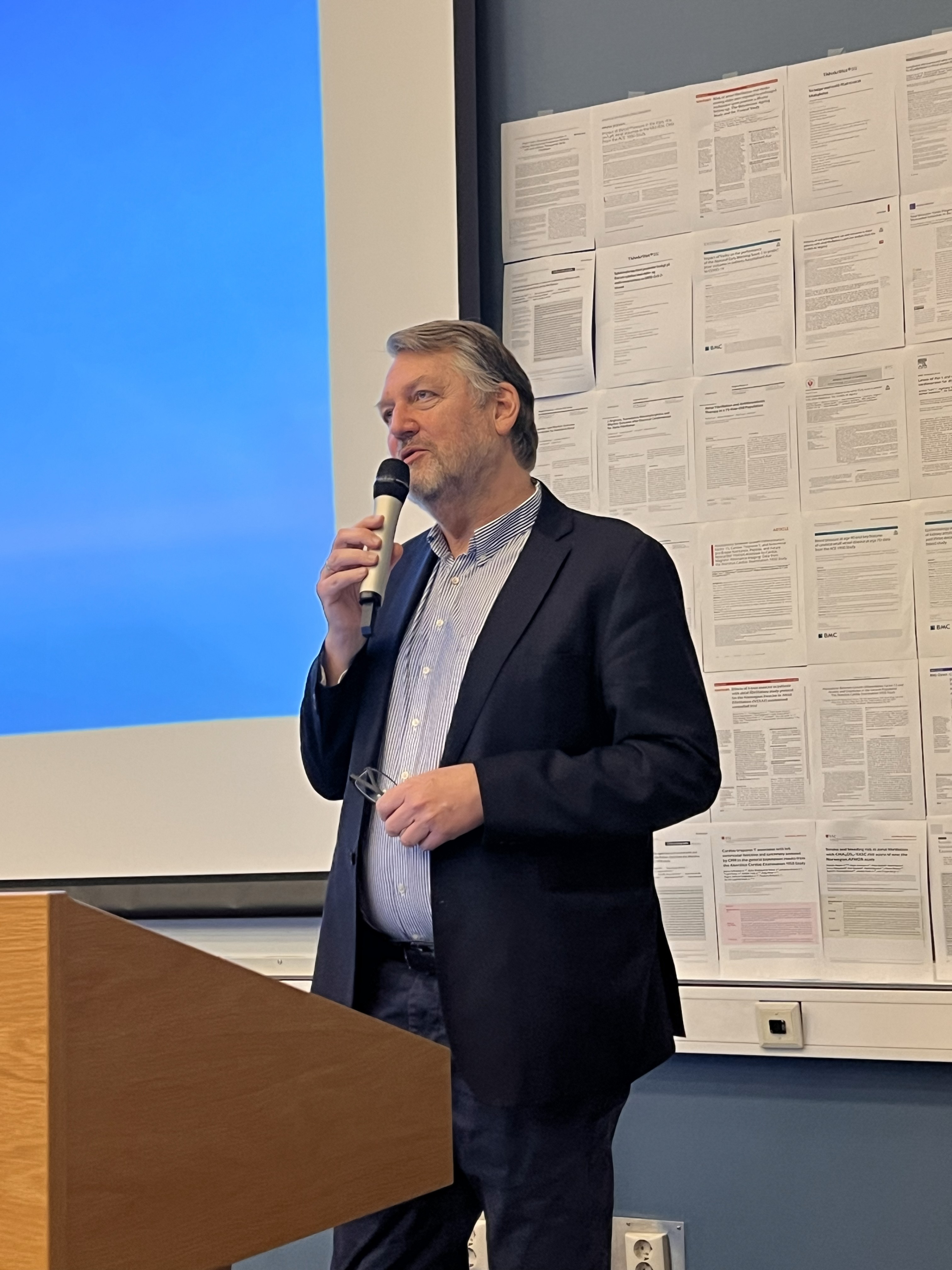 Alternativ tekst generert av copilot: En foredragsholder står ved talerstolen med mikrofon i hånden. I bakgrunnen er det en blå presentasjon og en vegg av opphengte vitenskapelige artikler som gir rommet et tydelig forskningspreg.
