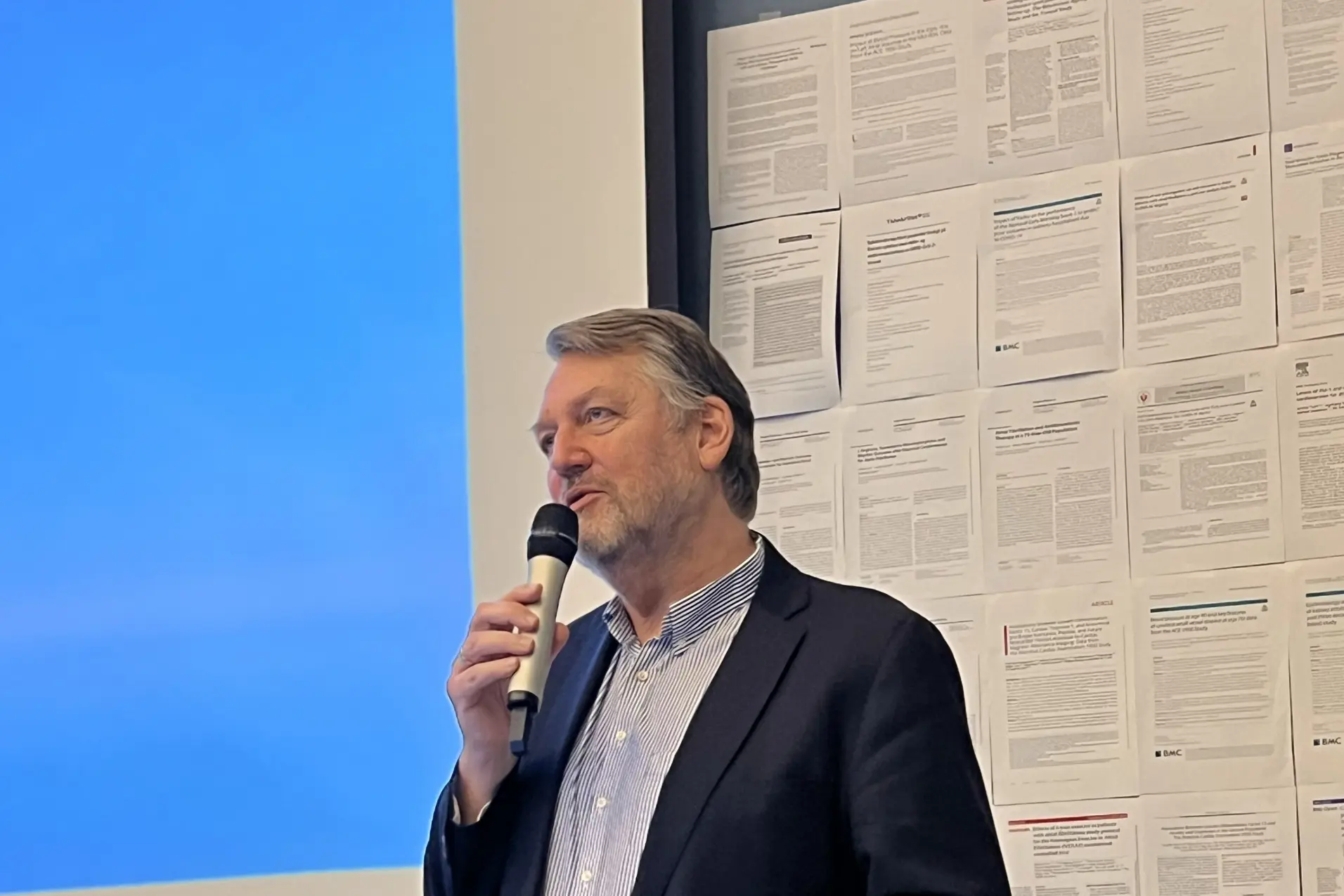 Alternativ tekst generert av copilot: En foredragsholder står ved talerstolen med mikrofon i hånden. I bakgrunnen er det en blå presentasjon og en vegg av opphengte vitenskapelige artikler som gir rommet et tydelig forskningspreg.