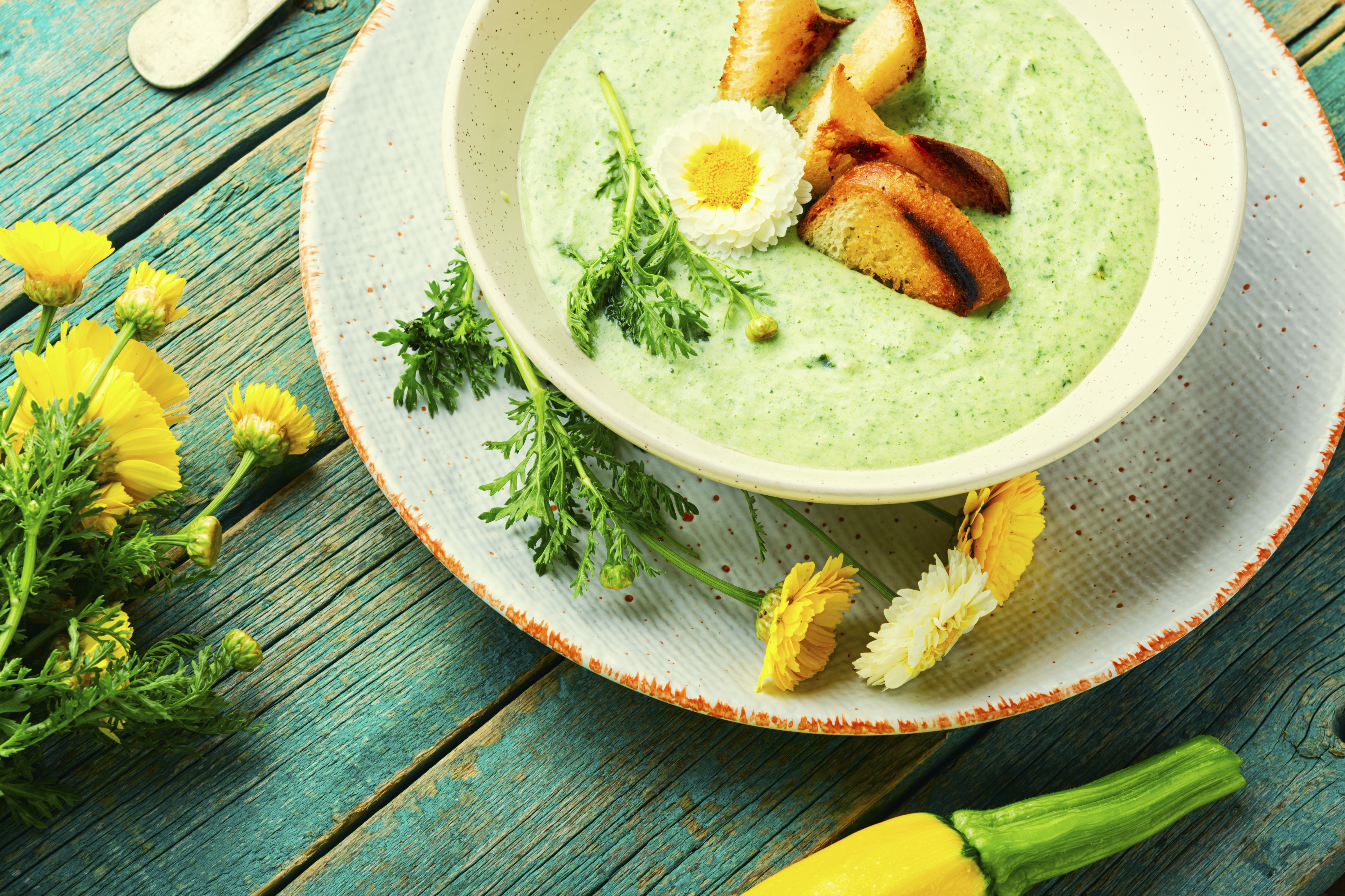 En bolle med grønn suppe toppet med krutonger og en spiselig blomst står på en tallerken omgitt av friske gule blomster på et blått trebord