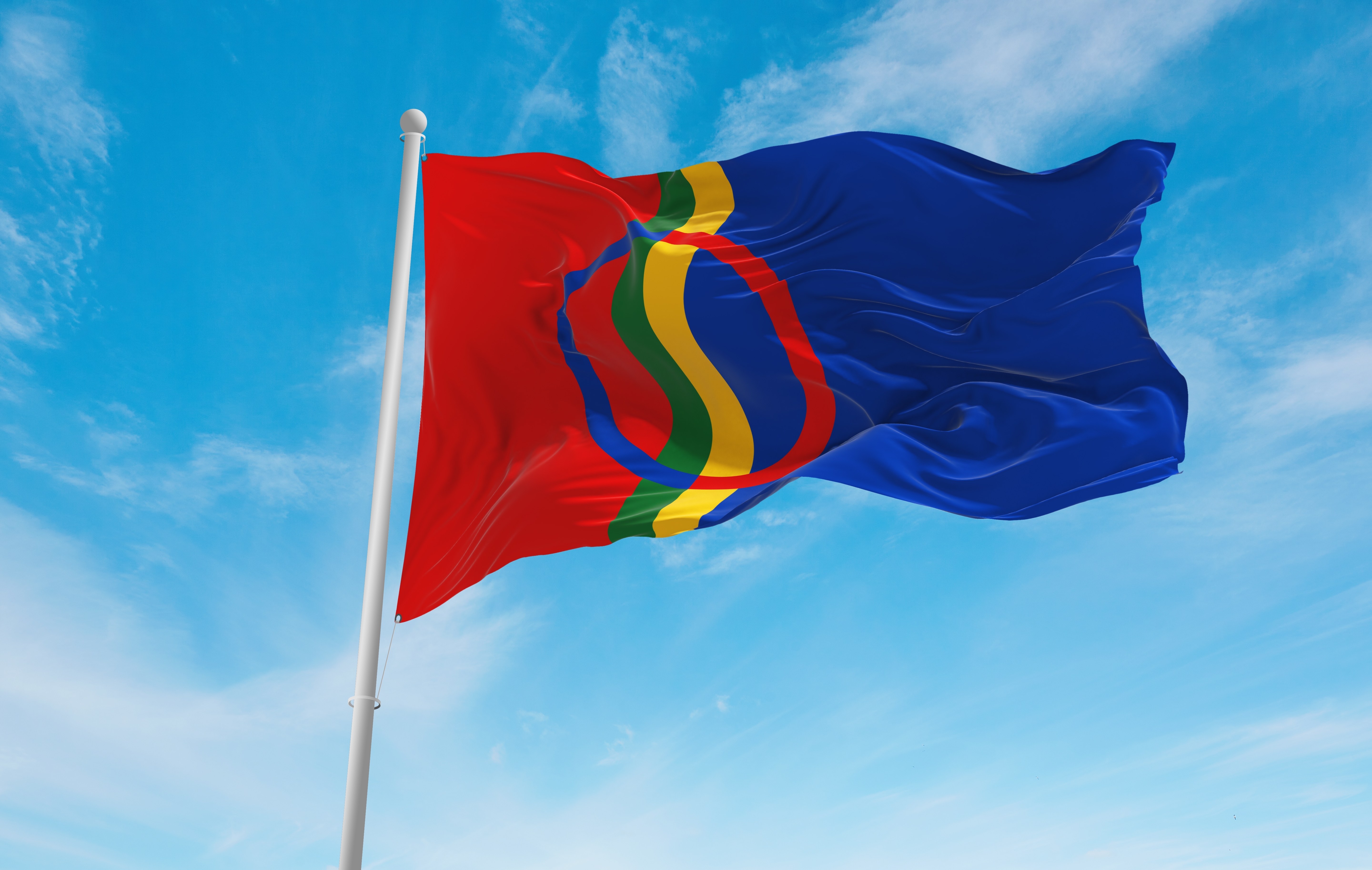 Samiske flagget som veiver i vinden på en flaggstang. Blå himmel med svake skyer bak.