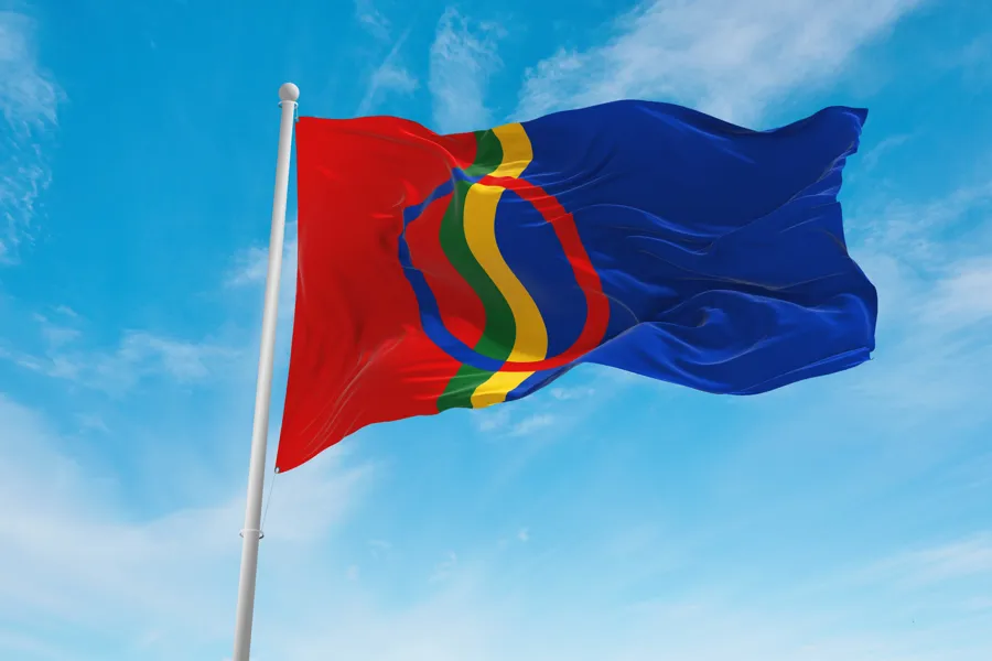 Samiske flagget som veiver i vinden på en flaggstang. Blå himmel med svake skyer bak.