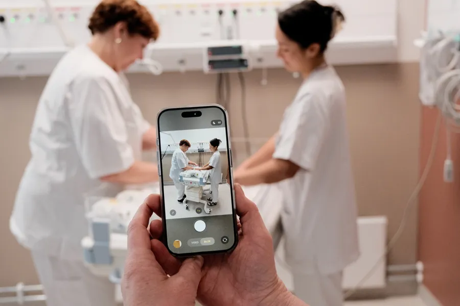 En person holder en mobiltelefon som viser et bilde av helsepersonell som arbeider rundt en kuvøse på et behandlingsrom.