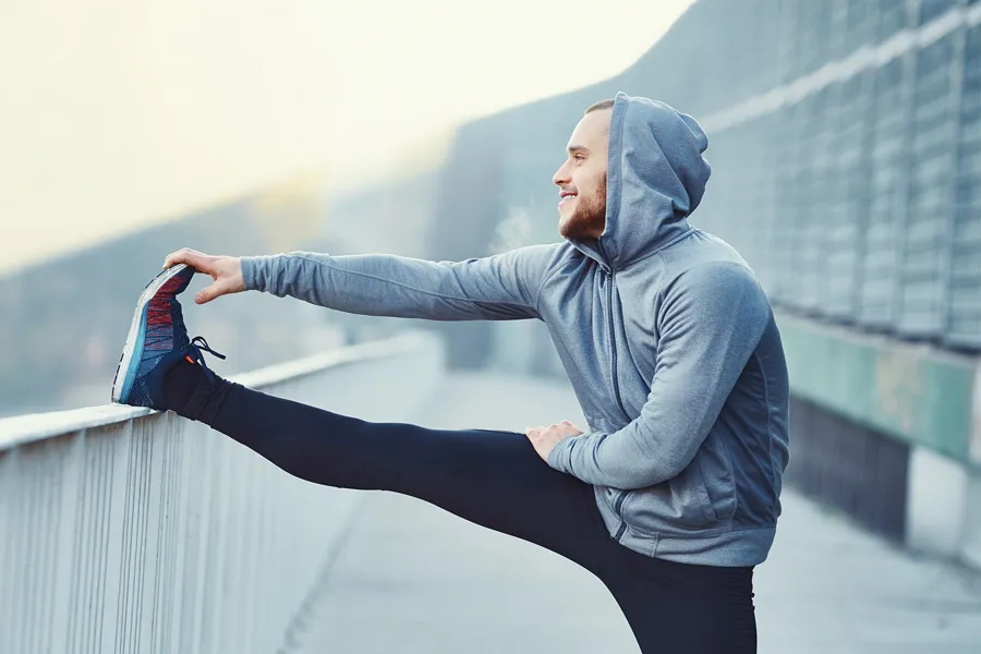 En mann i grå hettegenser, treningstights og joggesko som tøyer ene beinet på et rekkverk.