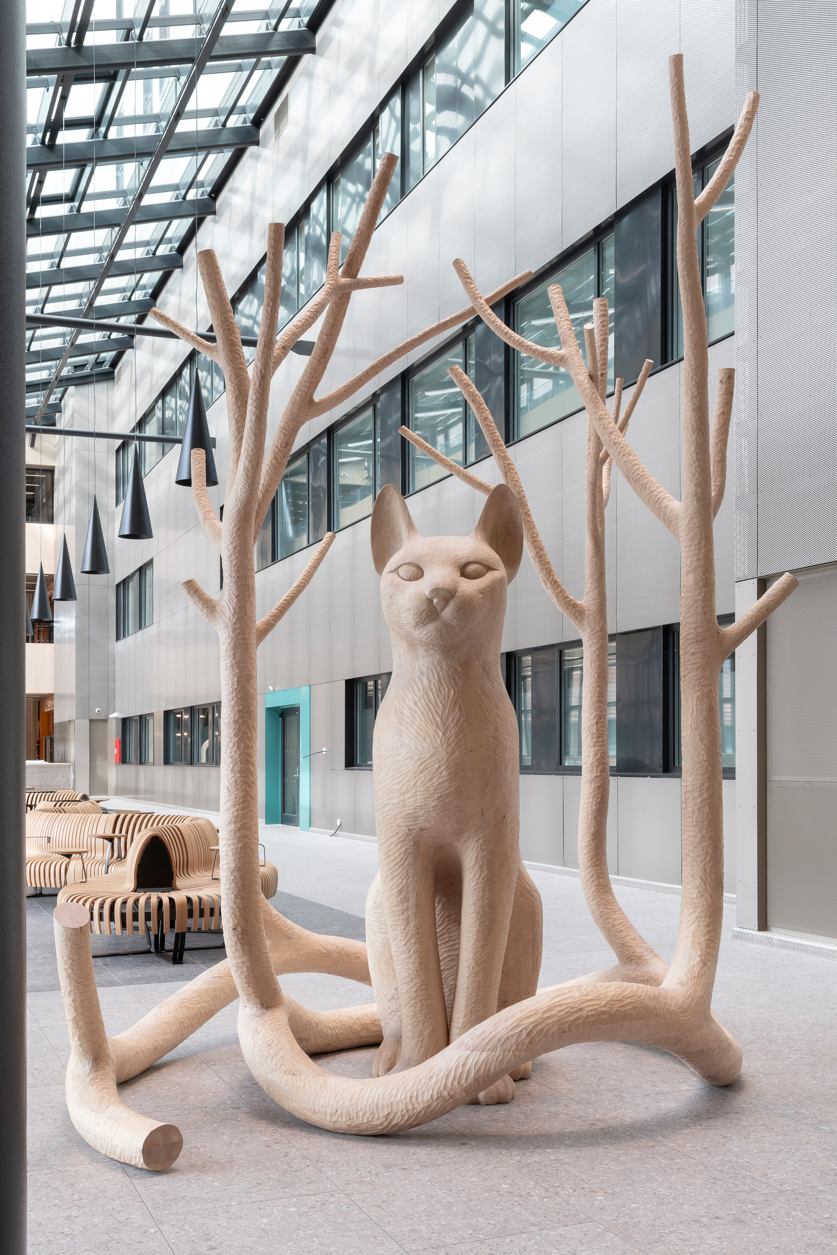 Ein stor katt sit majestetisk og ventar. Ut av halen har det grodd fire tre som omsluttar katten. Greinene er avskorne. Trea og halen lagar eit rom ein kan gå inn i.