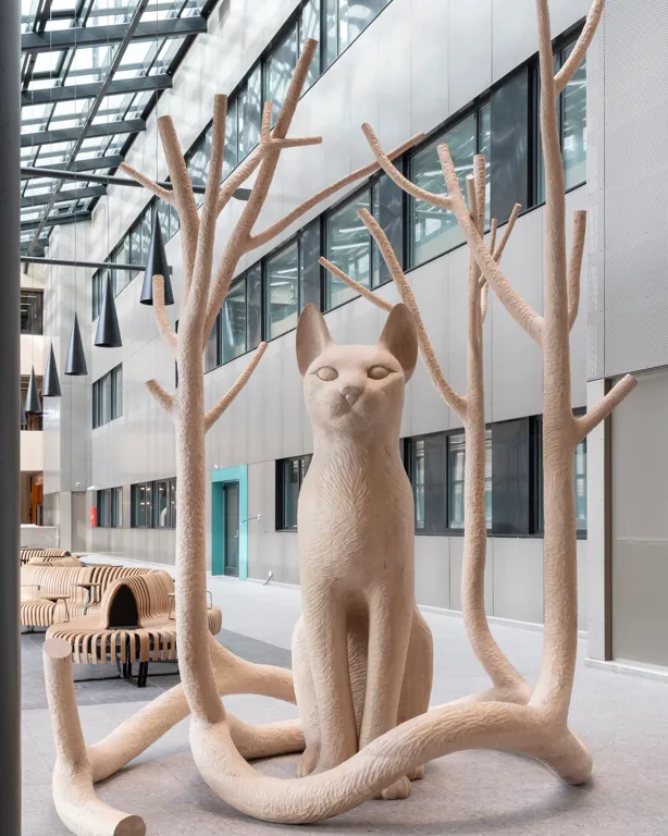 Ein stor katt sit majestetisk og ventar. Ut av halen har det grodd fire tre som omsluttar katten. Greinene er avskorne. Trea og halen lagar eit rom ein kan gå inn i.