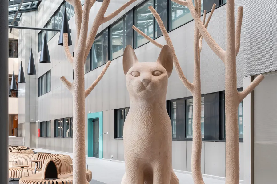 Ein stor katt sit majestetisk og ventar. Ut av halen har det grodd fire tre som omsluttar katten. Greinene er avskorne. Trea og halen lagar eit rom ein kan gå inn i.