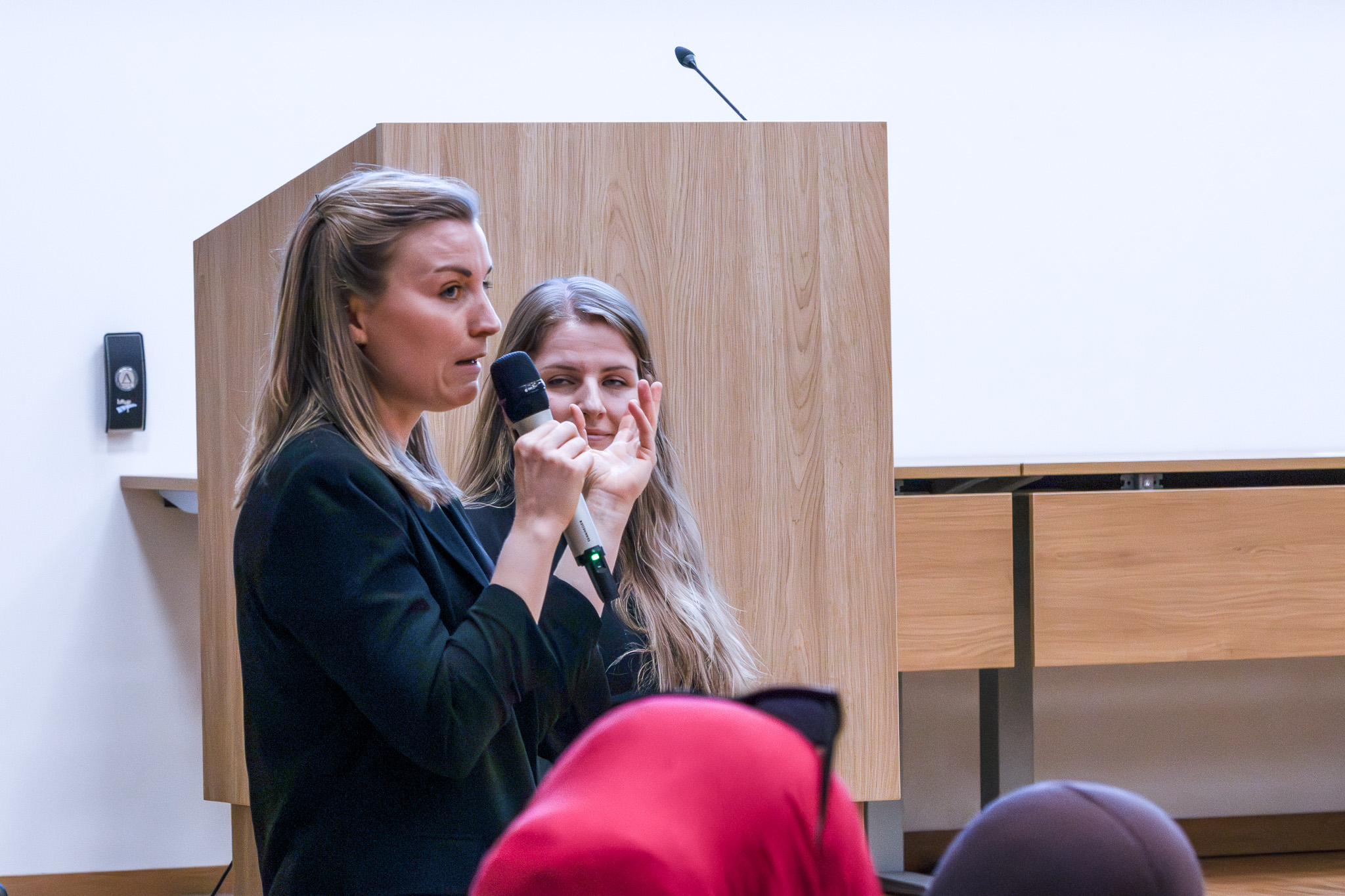 To personer står foran et podium, hvor den ene holder en mikrofon og snakker til publikum.