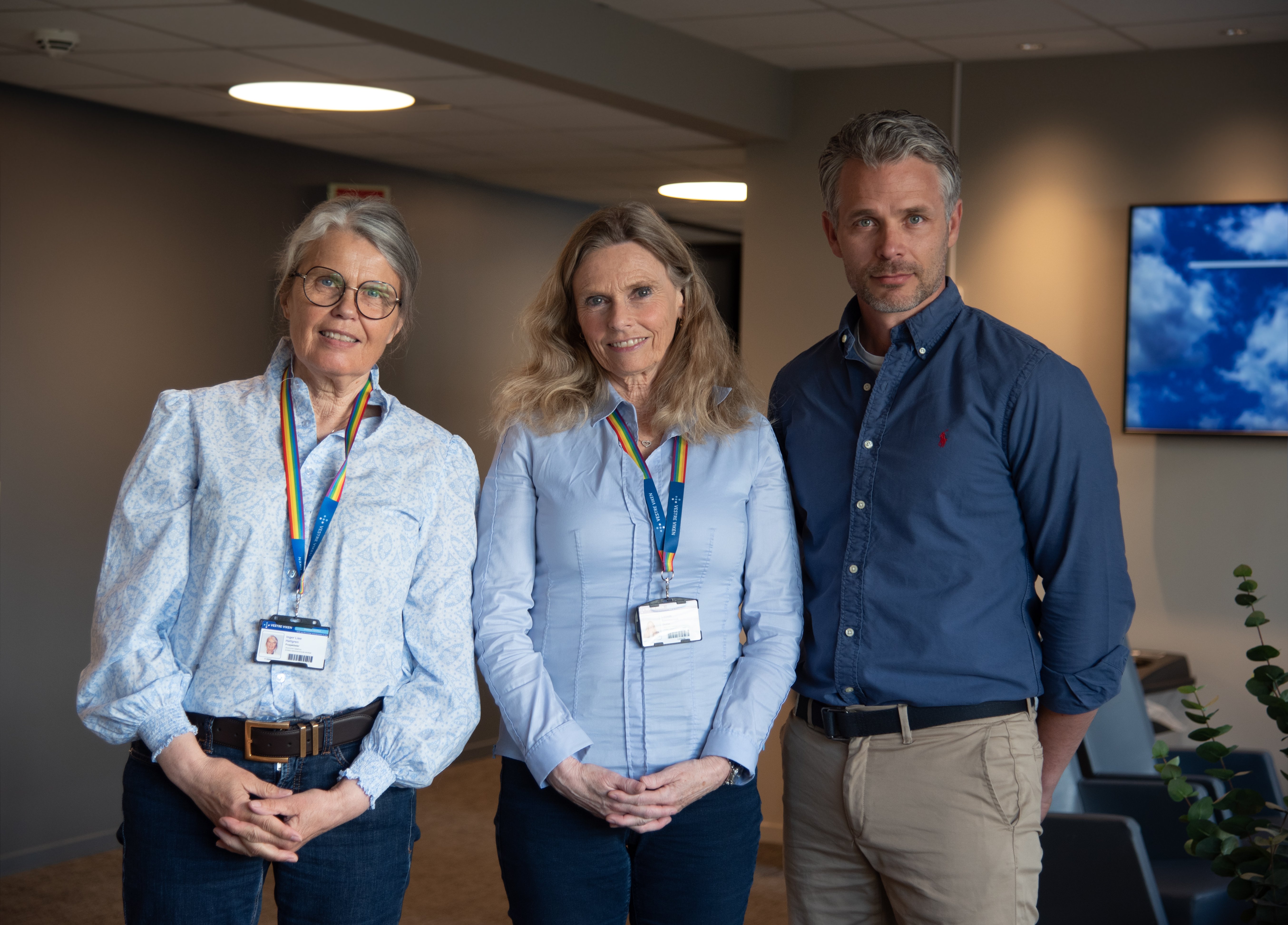 Tre personer som står oppstilt og ser mot kamera. To kvinner til venstre med lyseblå skjorte og id-kort i bånd i regnbuens farger rundt halsen. Mann i mørkeblå skjorte tiø høyre. De står i et kontorlandskap/korridor med stoler og en tv-skjerm på veggen bak seg.