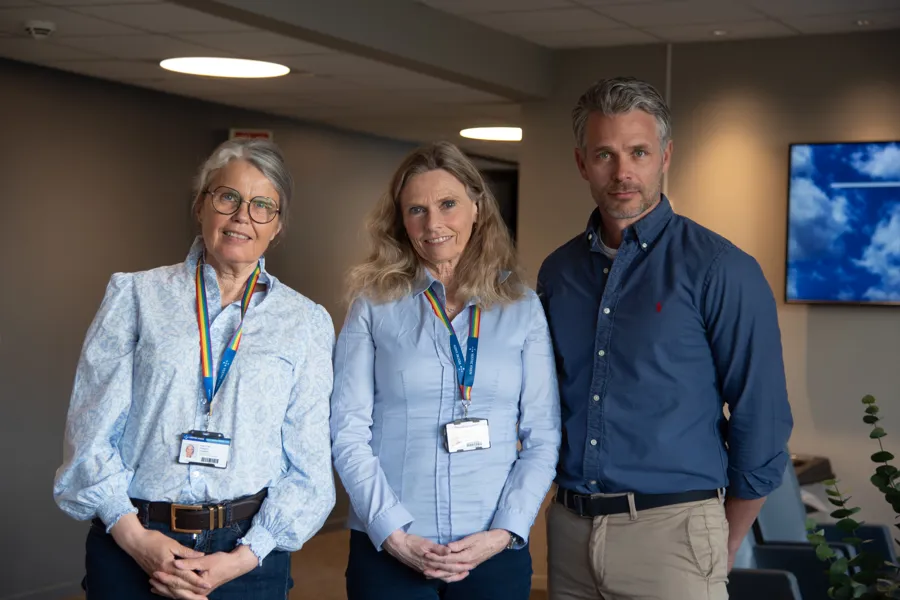Tre personer som står oppstilt og ser mot kamera. To kvinner til venstre med lyseblå skjorte og id-kort i bånd i regnbuens farger rundt halsen. Mann i mørkeblå skjorte tiø høyre. De står i et kontorlandskap/korridor med stoler og en tv-skjerm på veggen bak seg.