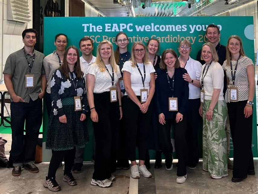 En gruppe mennesker som poserer for et bilde. De står foran en stor poster med teksten "The EAPC welcomes you" og alle har id-kort som henger i et bånd rundt halsen.