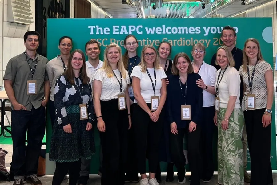 En gruppe mennesker som poserer for et bilde. De står foran en stor poster med teksten "The EAPC welcomes you" og alle har id-kort som henger i et bånd rundt halsen.