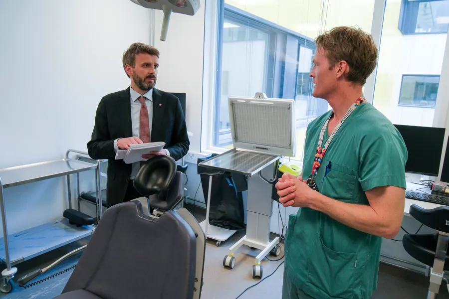 To personer i et behandlingsrom på en sykehusavdeling snakker sammen ved en undersøkelsesstol med medisinsk utstyr rundt.