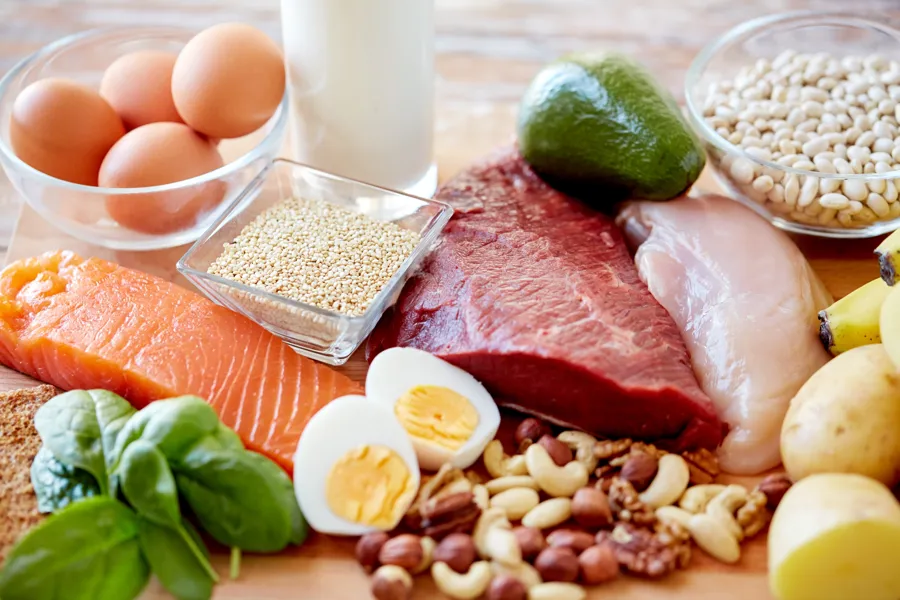 Råvarer av mat som ligger på et bord. Egg i en glasskål, melk i et glass, rødt kjøtt, kyllingfillet, laksefillet, kokt egg delt i to, diverse nøtter, basilikumblader, poteter delt i to, hvite tørkede bønner i glasskål og bananer
