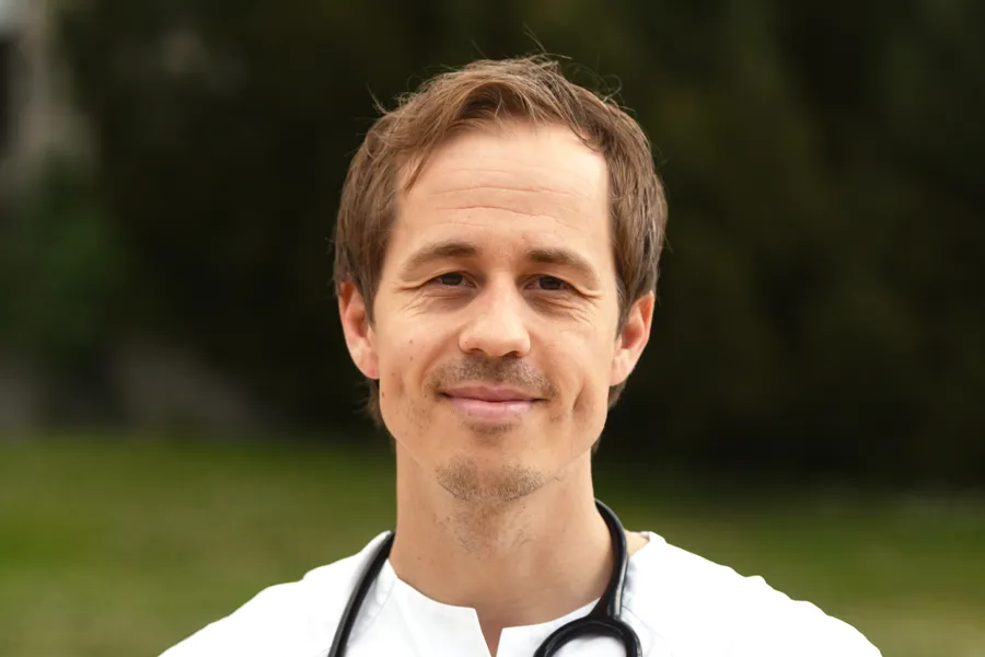 Portrett av en lege ute i naturen iført sykehusuniform. Han har stetoskop rundt halsen og ID-kort og penn i brystlommen.