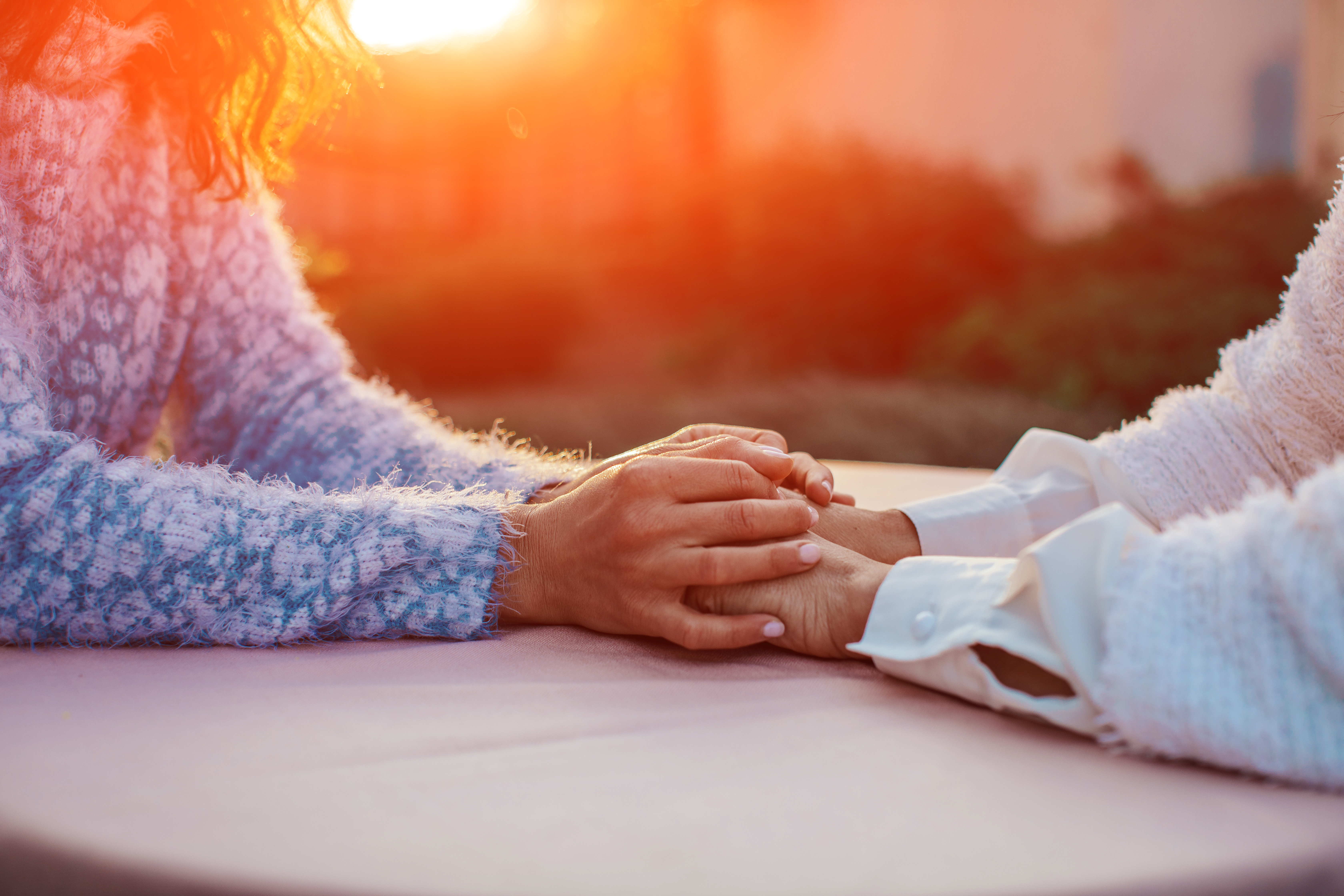 En person som holder hendene oppå hendene til en annen person. De har armene/hendene på et bord og solen stråler bak personene