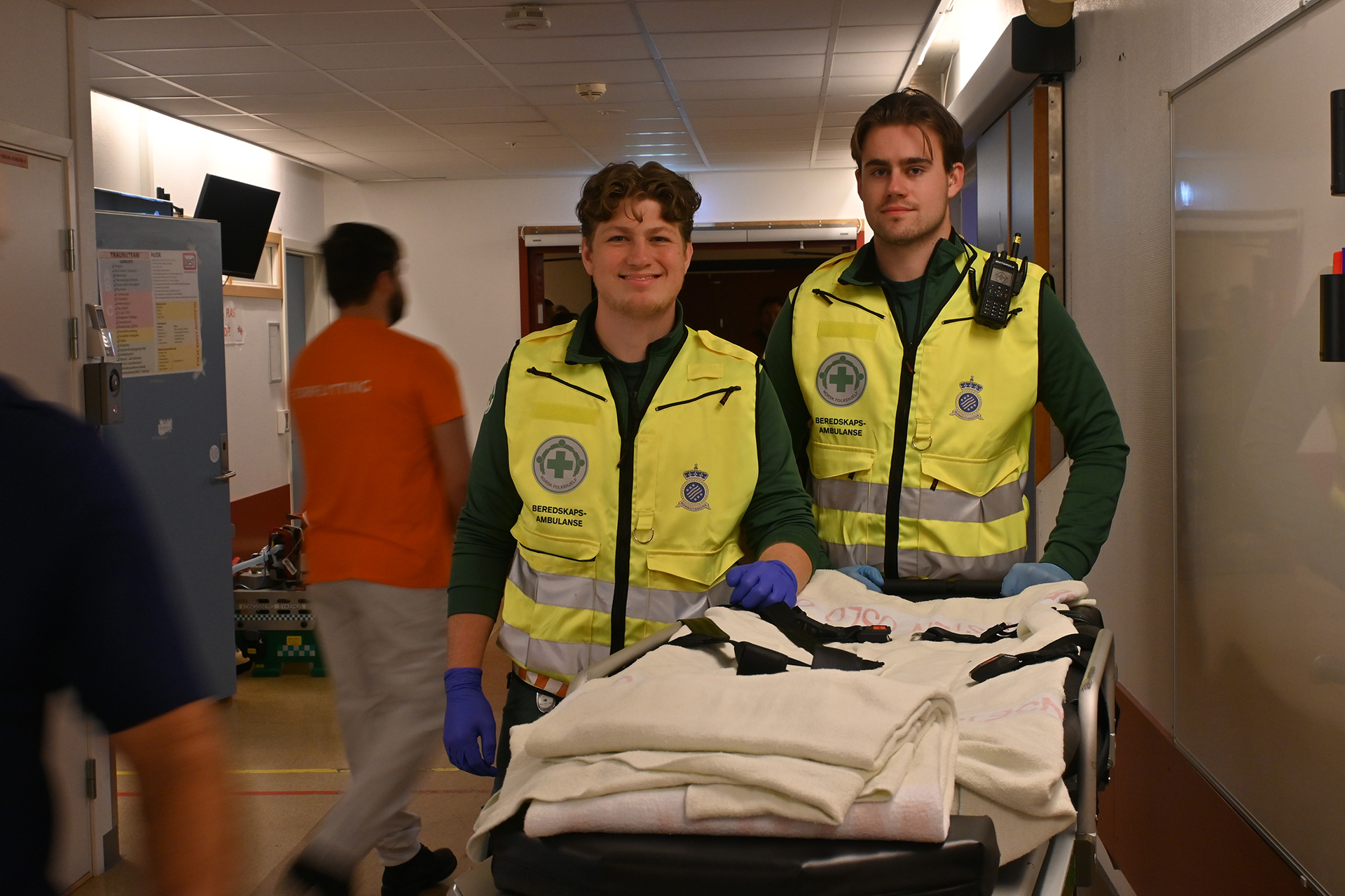 Et par menn i sikkerhetsvester som står ved siden av en seng