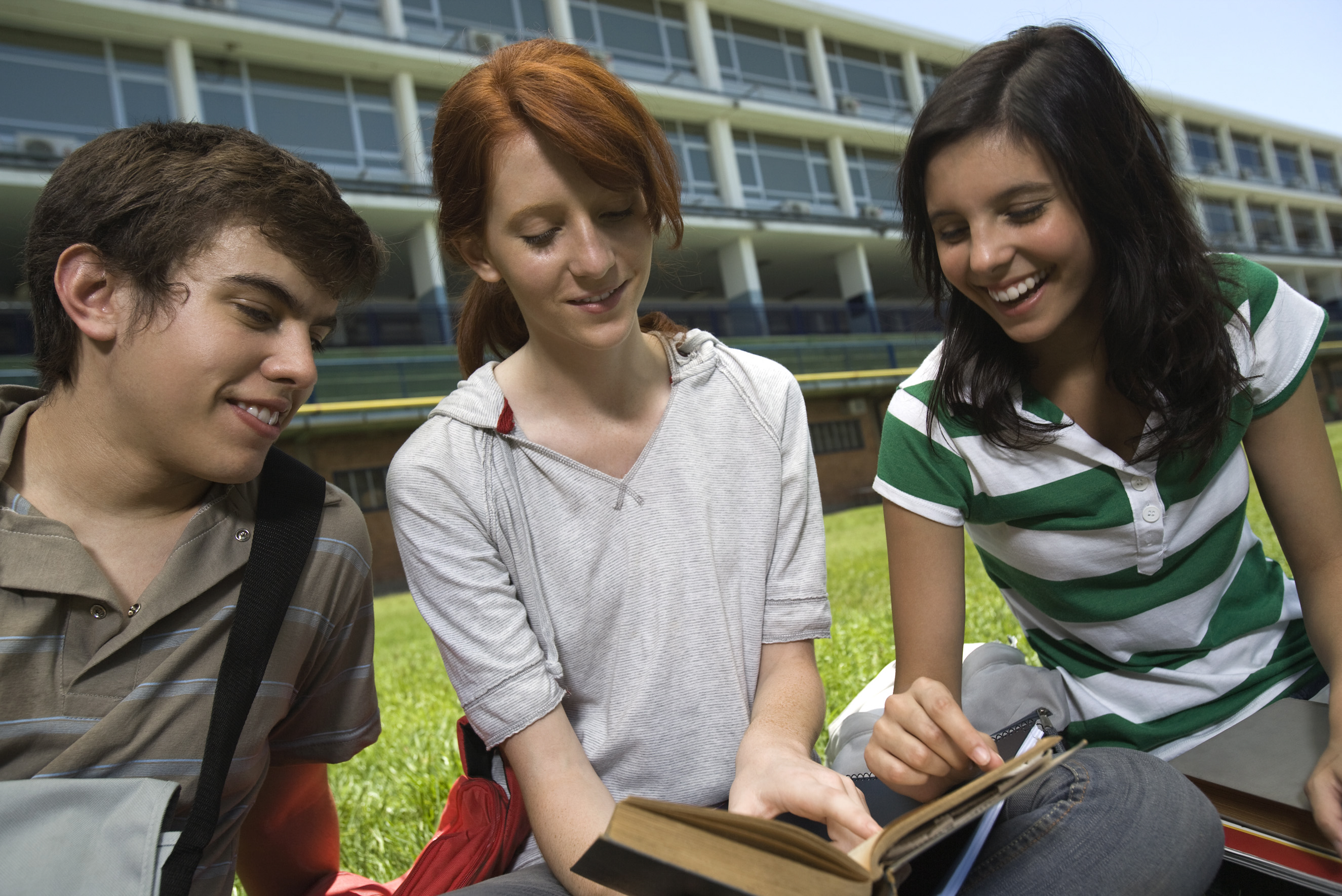 Tre ungdommer sitter på gressplenen utenfor en skole.