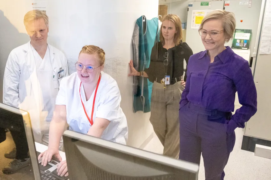 Fire personer i et sykehusmiljø står samlet rundt en dataskjerm. To ansatte i hvite uniformer ser på skjermen, mens to andre observatører bak glass følger med og smiler.