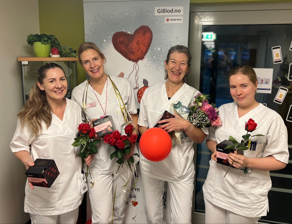 Fire personer i hvite arbeidsklær holder roser, konfektesker, blomster og en rød ballong foran en plakat med teksten ‘GiBlod.no’ og Røde Kors-logo