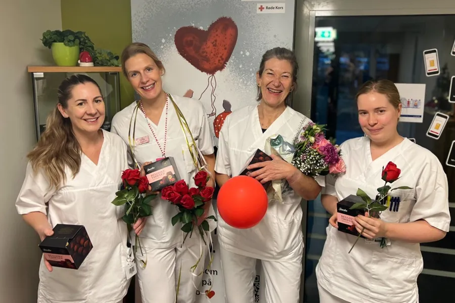 Fire personer i hvite arbeidsklær holder roser, konfektesker, blomster og en rød ballong foran en plakat med teksten ‘GiBlod.no’ og Røde Kors-logo