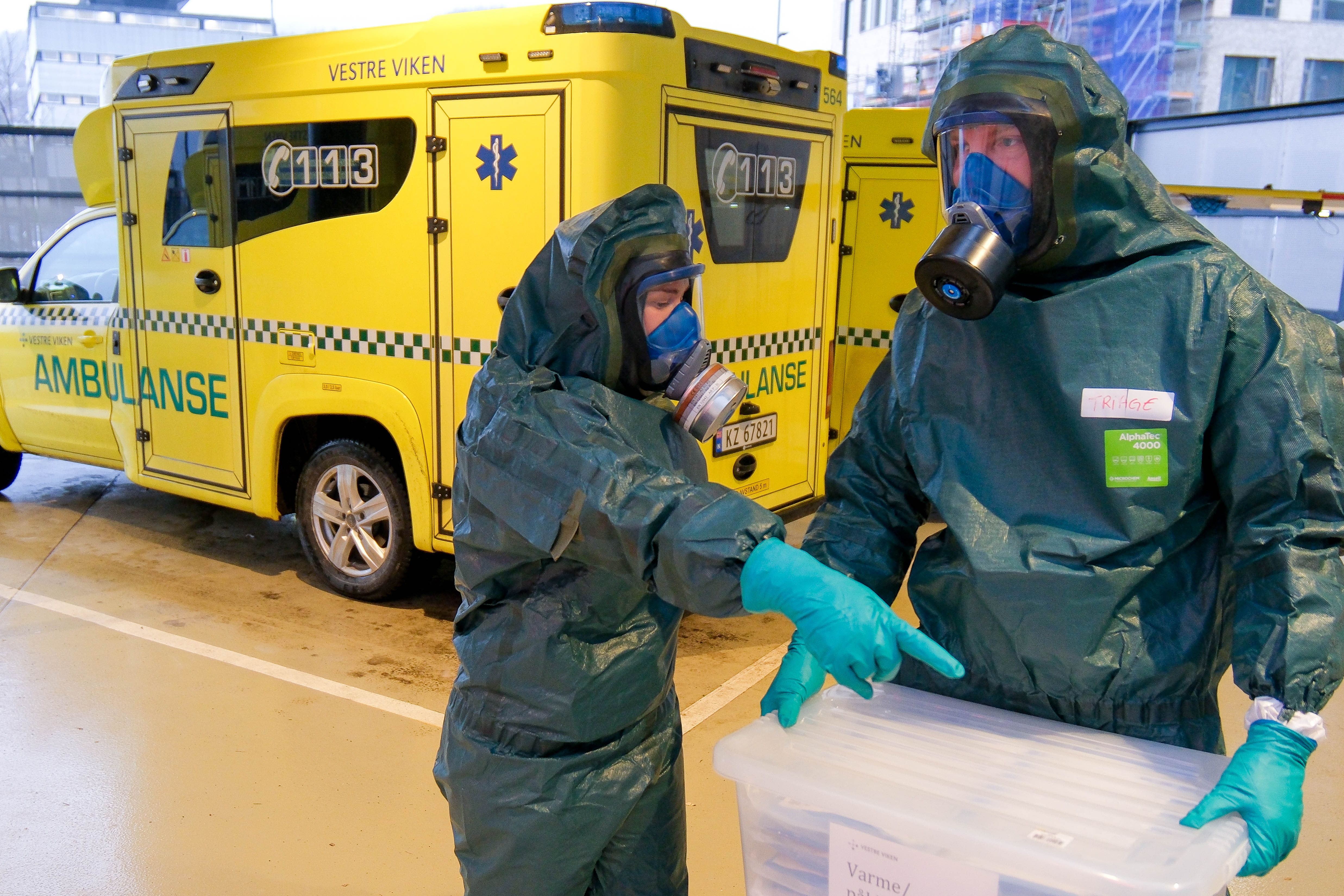 To personer i vernedrakt med gassmasker håndterer plastkasse foran gul ambulanse.