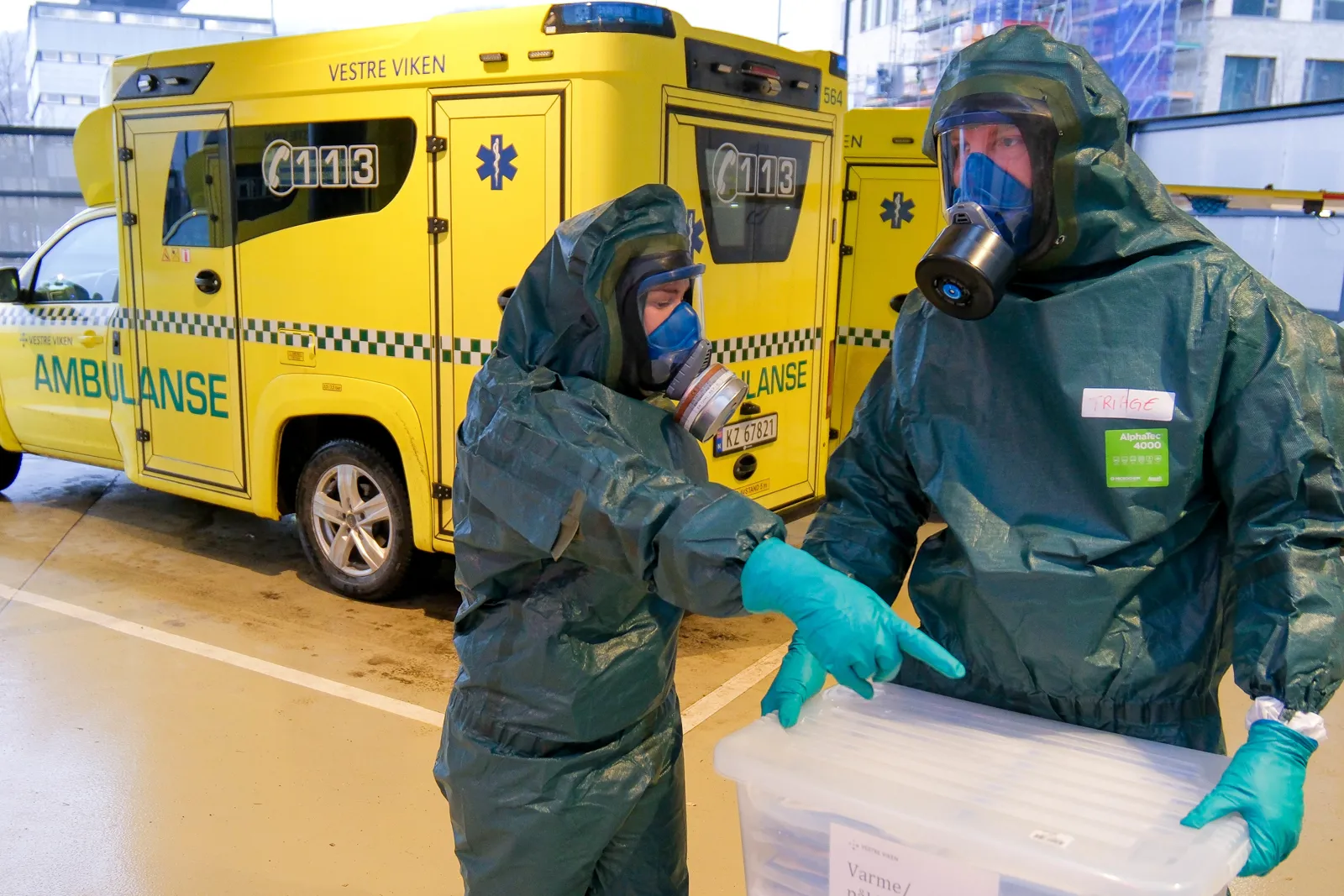 To personer i vernedrakt med gassmasker håndterer plastkasse foran gul ambulanse.