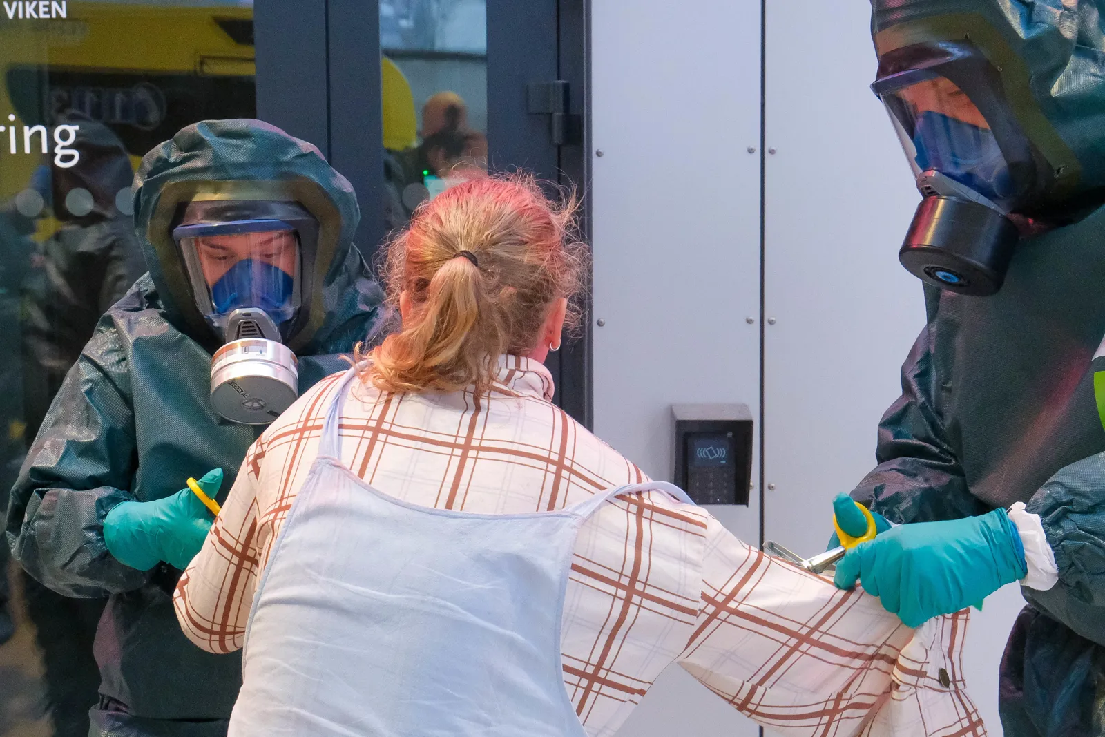 Personer i vernedrakt hjelper pasient med rutete skjorte ved inngangsparti på sykehuset.