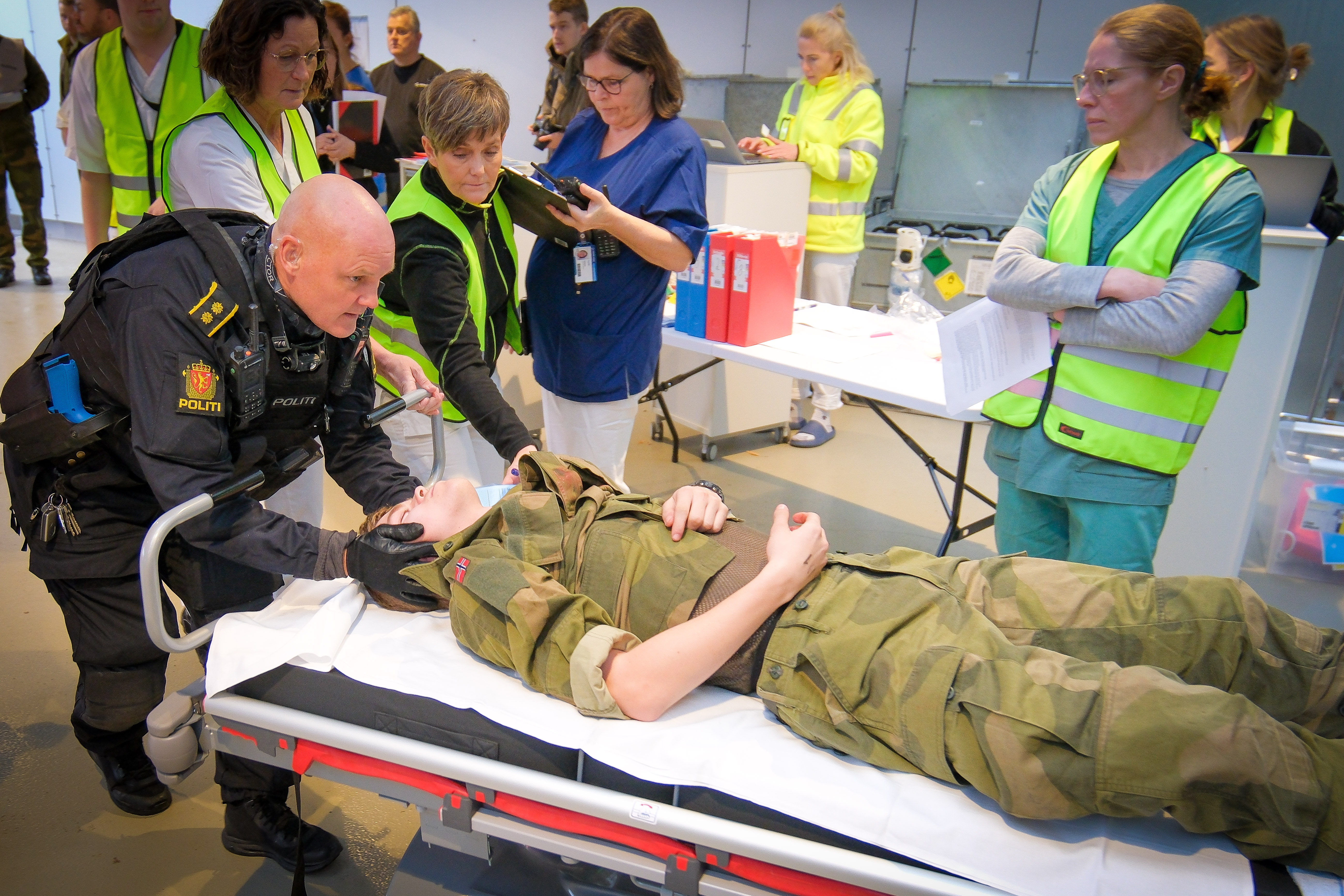 Politi bøyer seg over person i militæruniform på båre, flere personer i bakgrunnen.