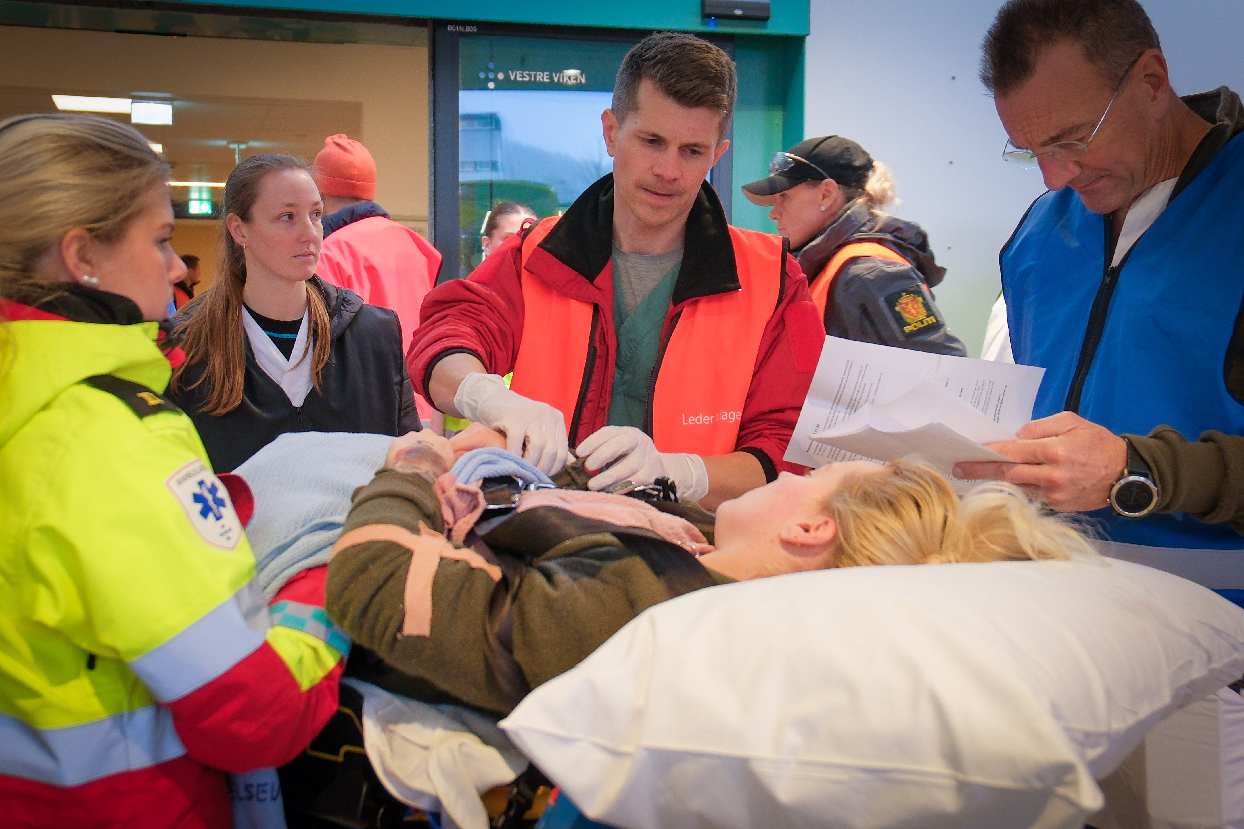 Person på båre får behandling av flere personer i fargerike vester under øvelse.