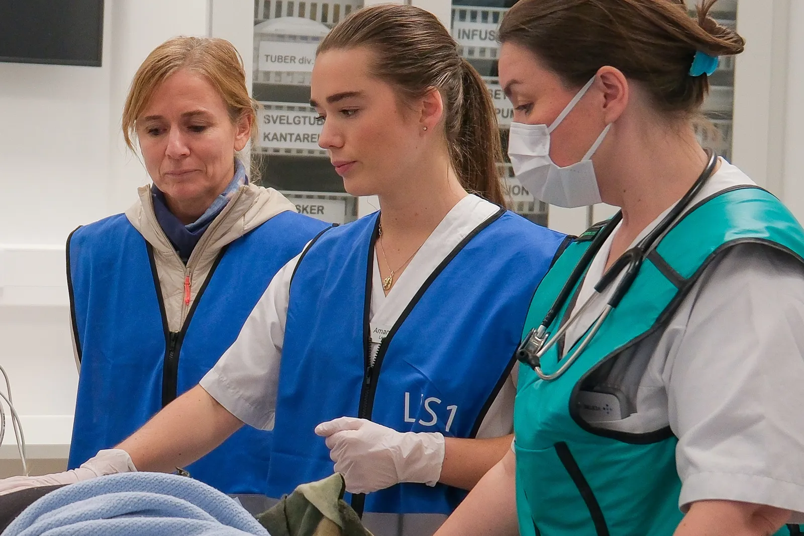 Tre personer med blå vester står ved pasient på båre i akuttmottak.