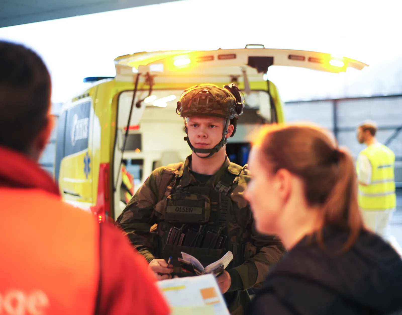 Militærperson med hjelm snakker med personer foran ambulanse med åpne dører.