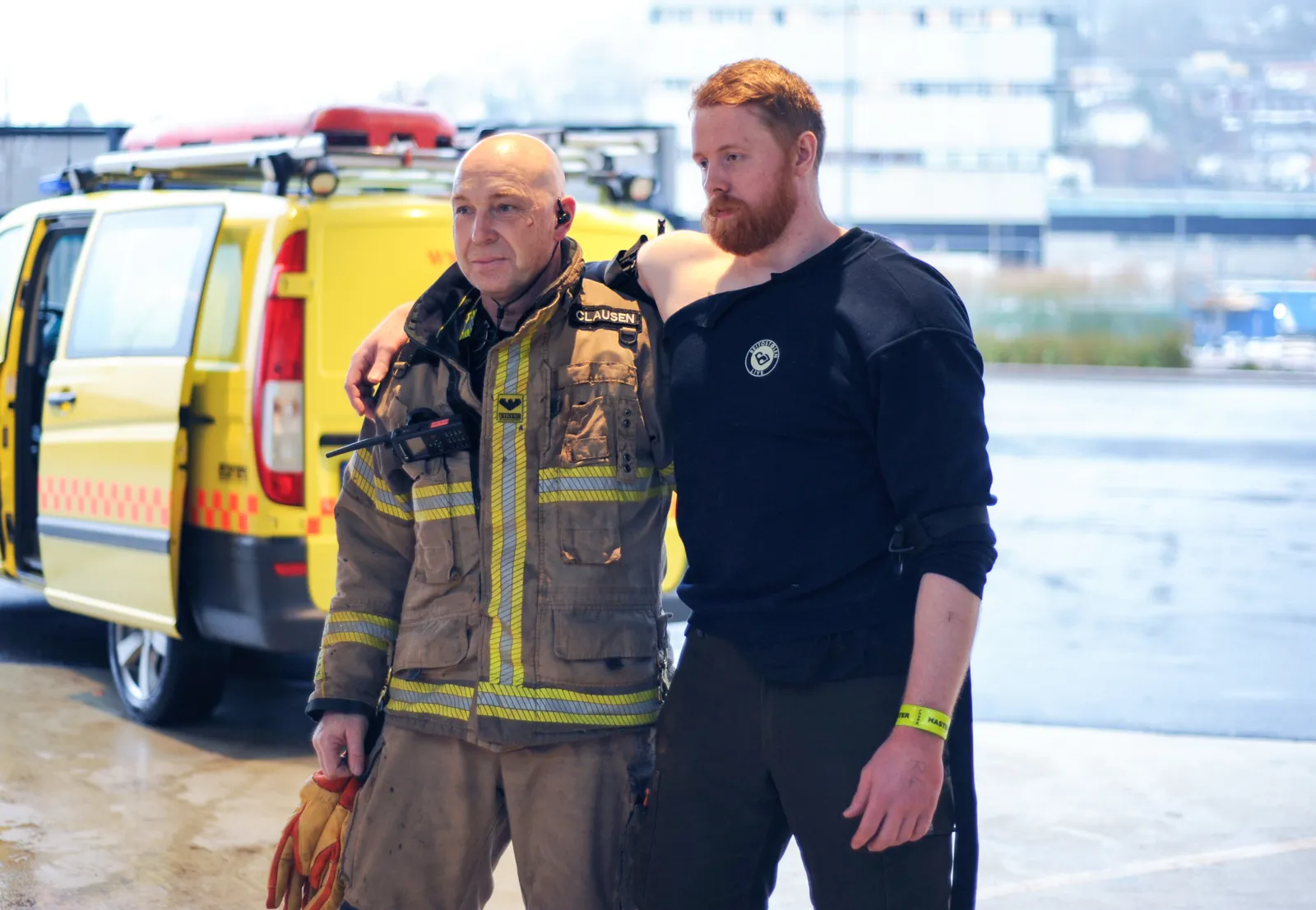 Brannmann i uniform står ved siden av person i mørke klær foran gul utrykningsbil.
