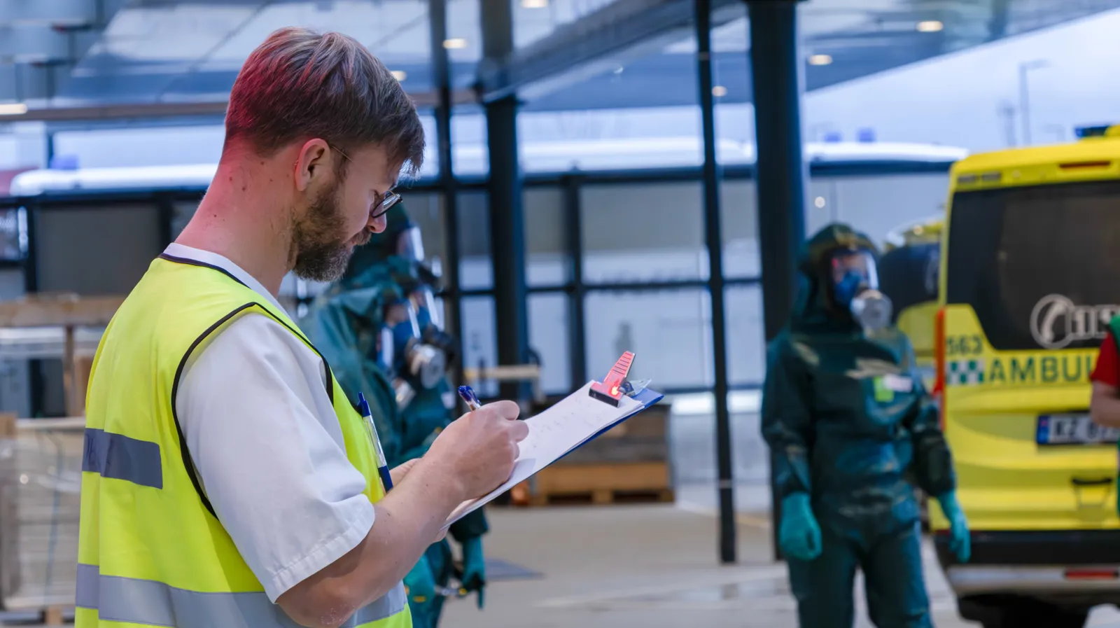 Person i gul vest skriver på sjekkliste mens to personer i vernedrakt står foran gul ambulanse.
