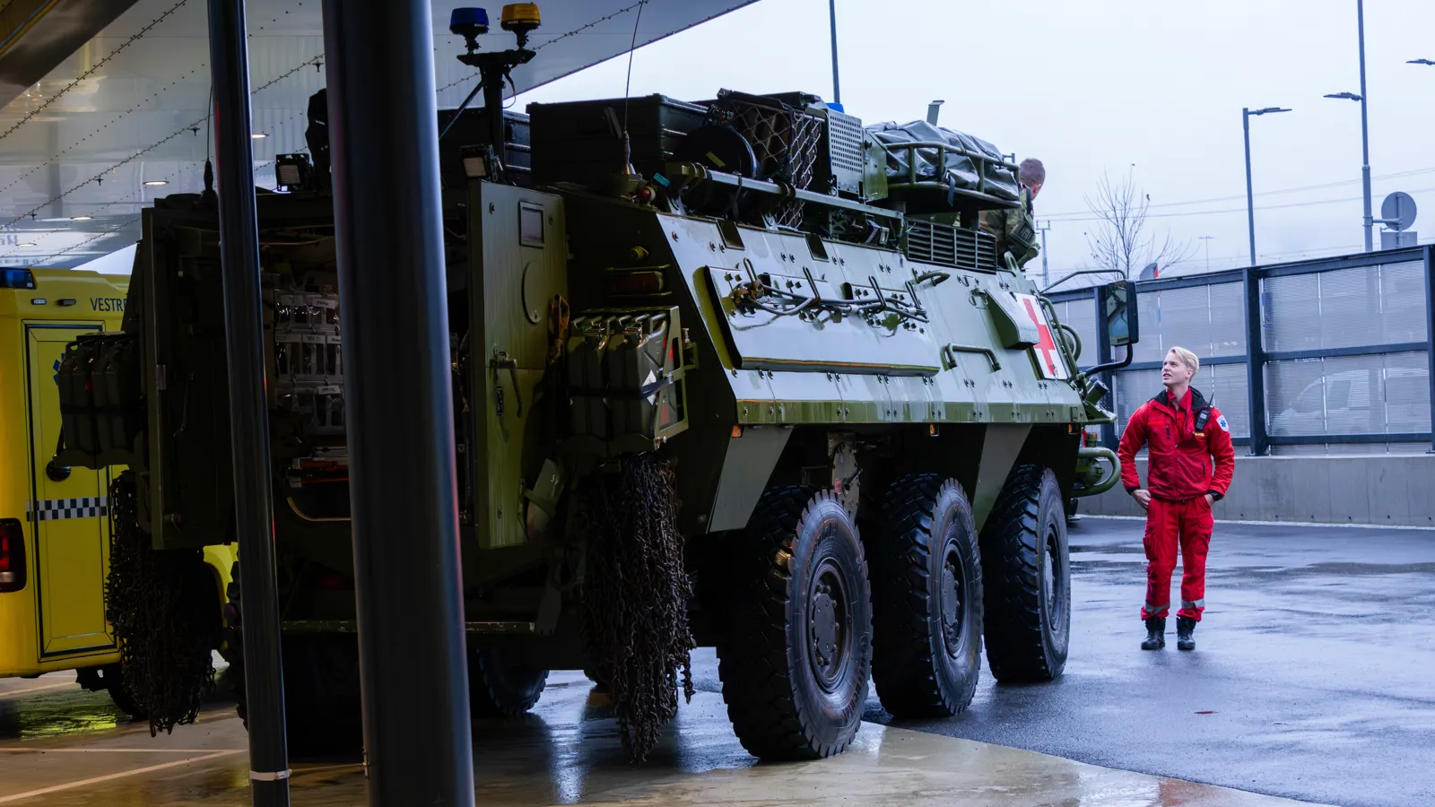 Militært kjøretøy med seks hjul står parkert ved ambulanse, person i rød drakt observerer.