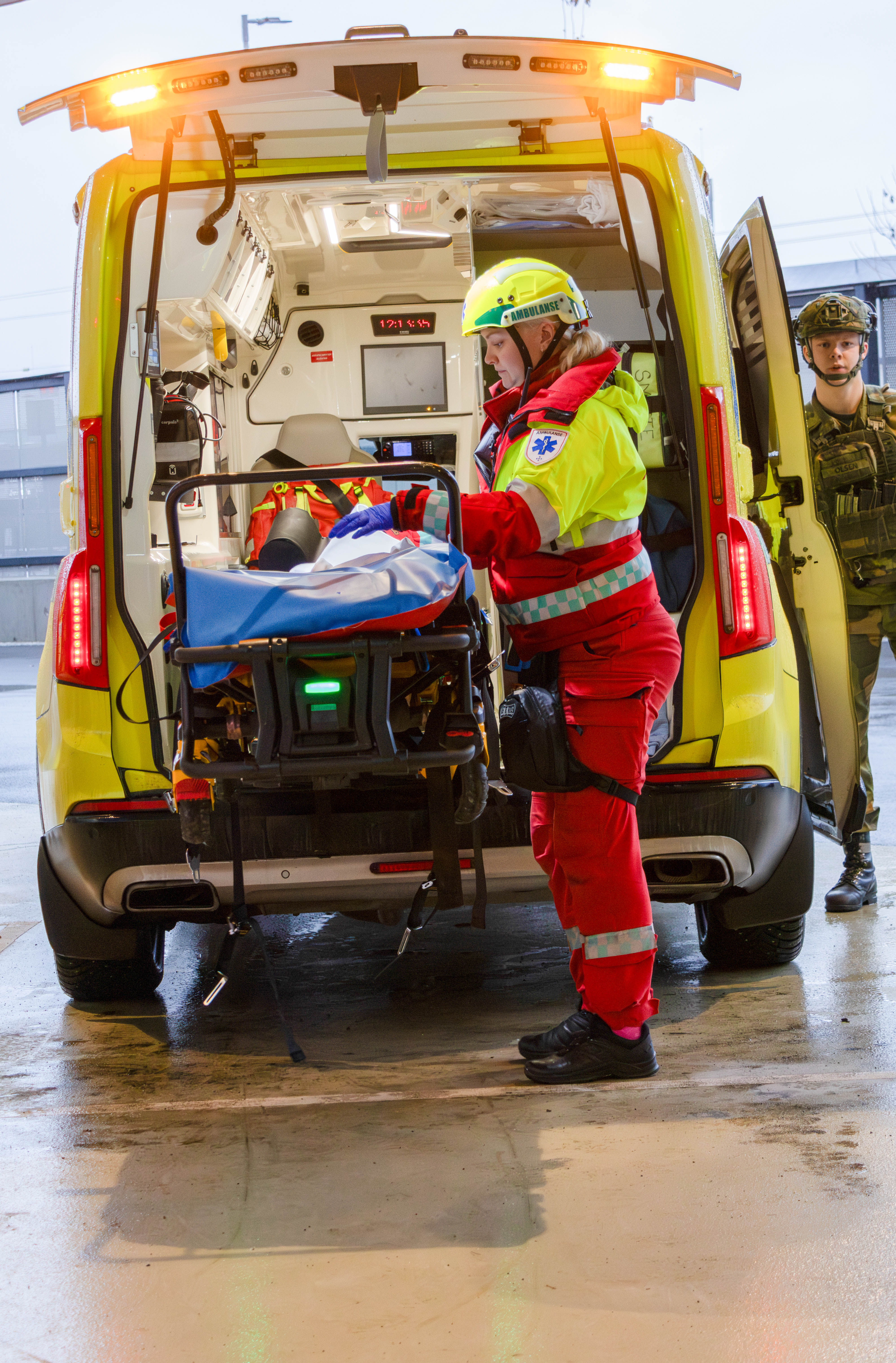 Ambulansepersonell i rød drakt laster båre med pasient inn i gul ambulanse, militært kjøretøy i bakgrunnen.