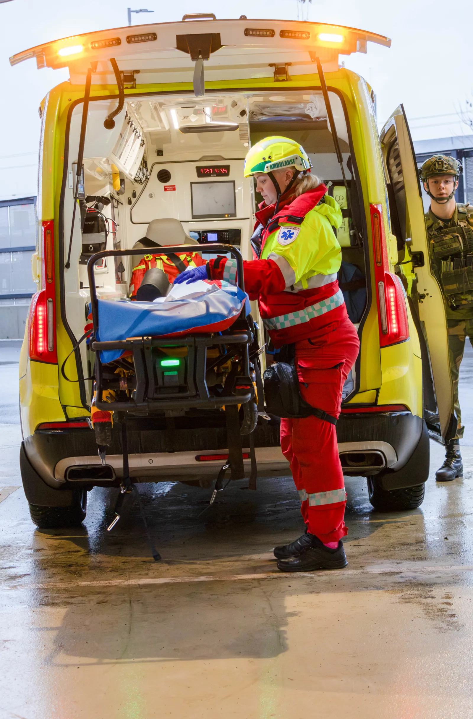 Ambulansepersonell i rød drakt laster båre med pasient inn i gul ambulanse, militært kjøretøy i bakgrunnen.