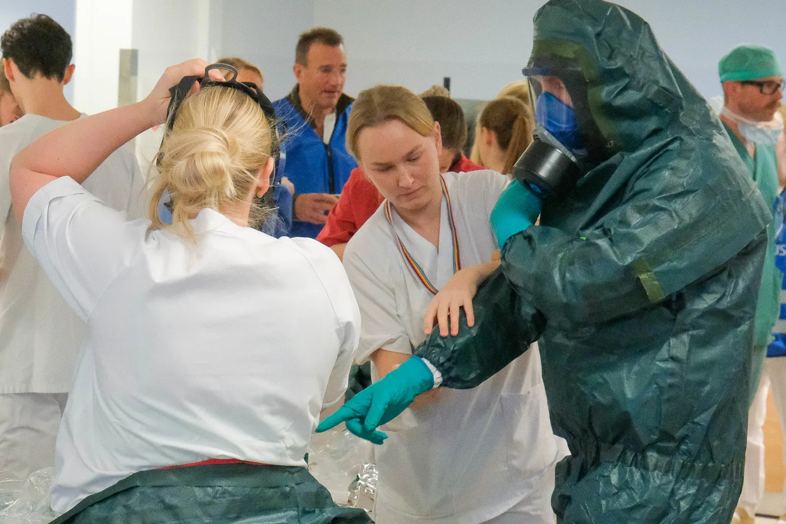 Person i vernedrakt med gassmaske gir instruksjoner til helsepersonell i hvite klær under øvelse.