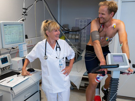 Bildet viser Turid Appeland som står ved siden av en pasient som sitter på en ergometersykkel